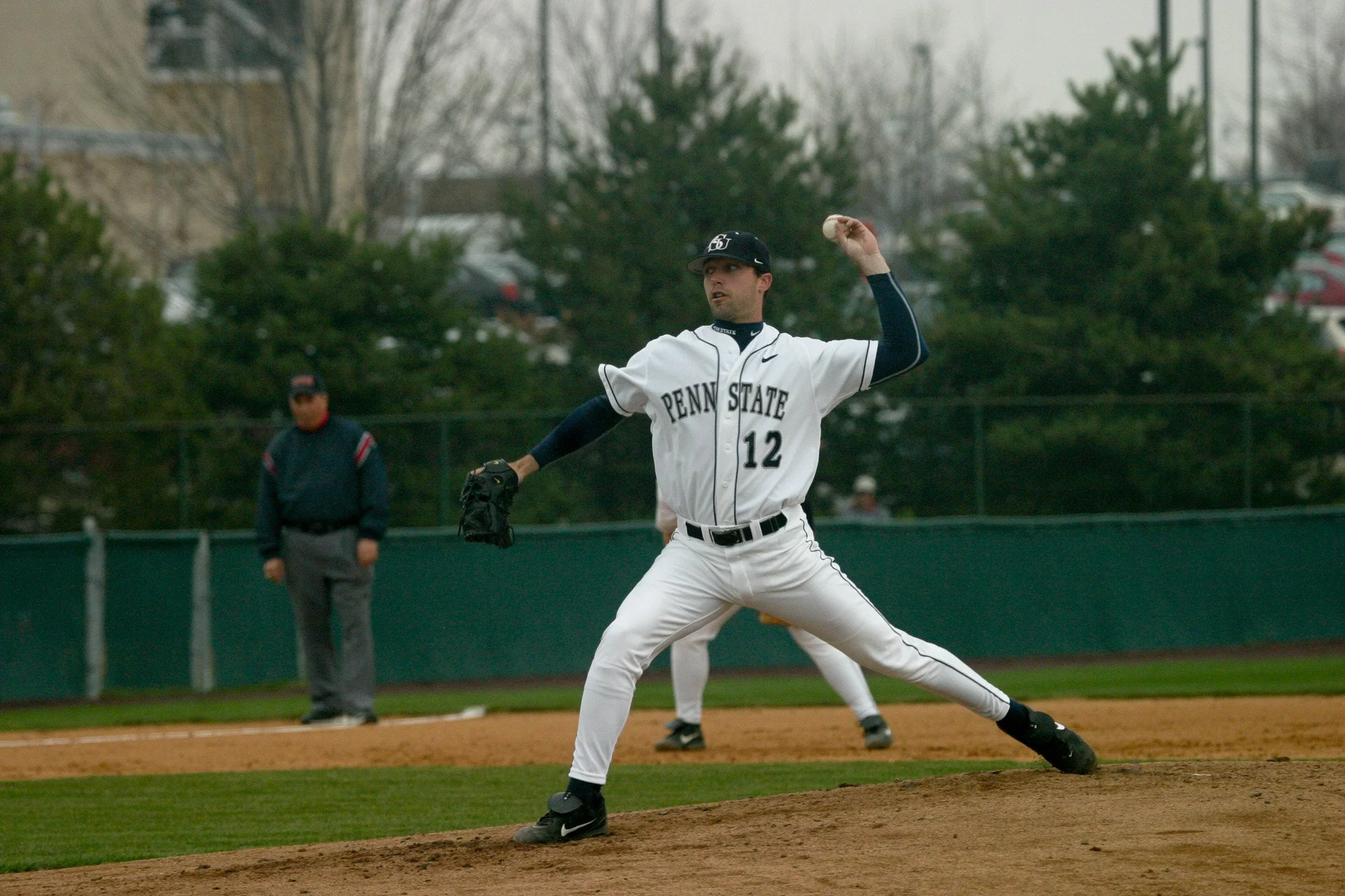 A baseball player from Penn State wearing a white uniform with the number 12 is pitching on the mound during a game. An umpire stands in the background, along with a blurred additional player.