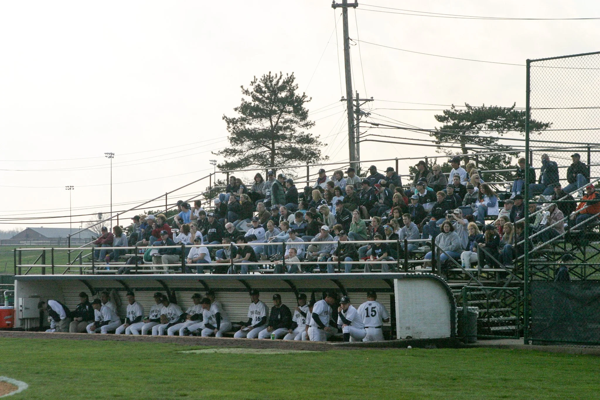Spectators sitting in bleachers at a baseball game, with players in the dugout in the foreground