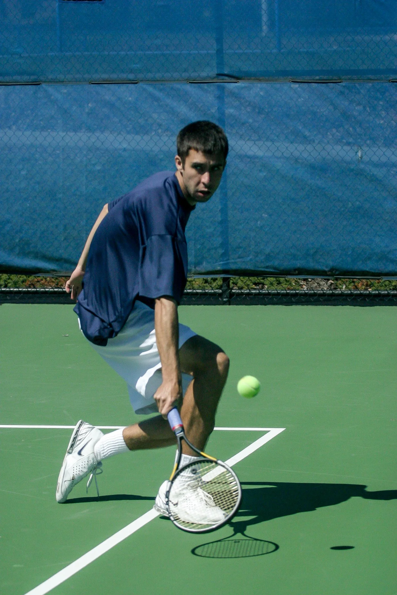 A male tennis player on a green tennis court, wearing a blue shirt, white shorts, and white shoes, preparing to hit a tennis ball with his racket.