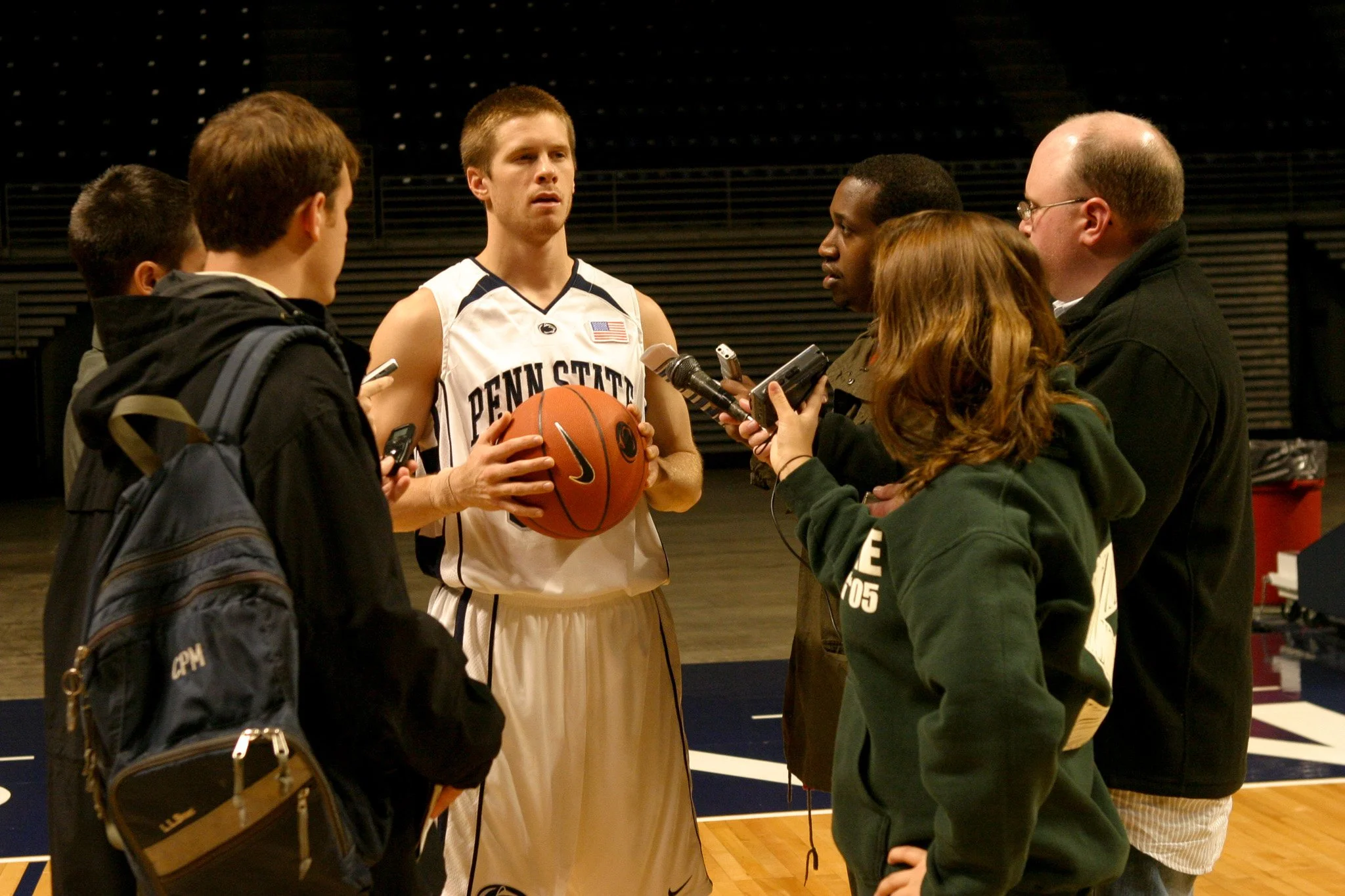A Penn State basketball player in uniform holding a basketball, surrounded by reporters and journalists with microphones and recording devices during a post-game interview.