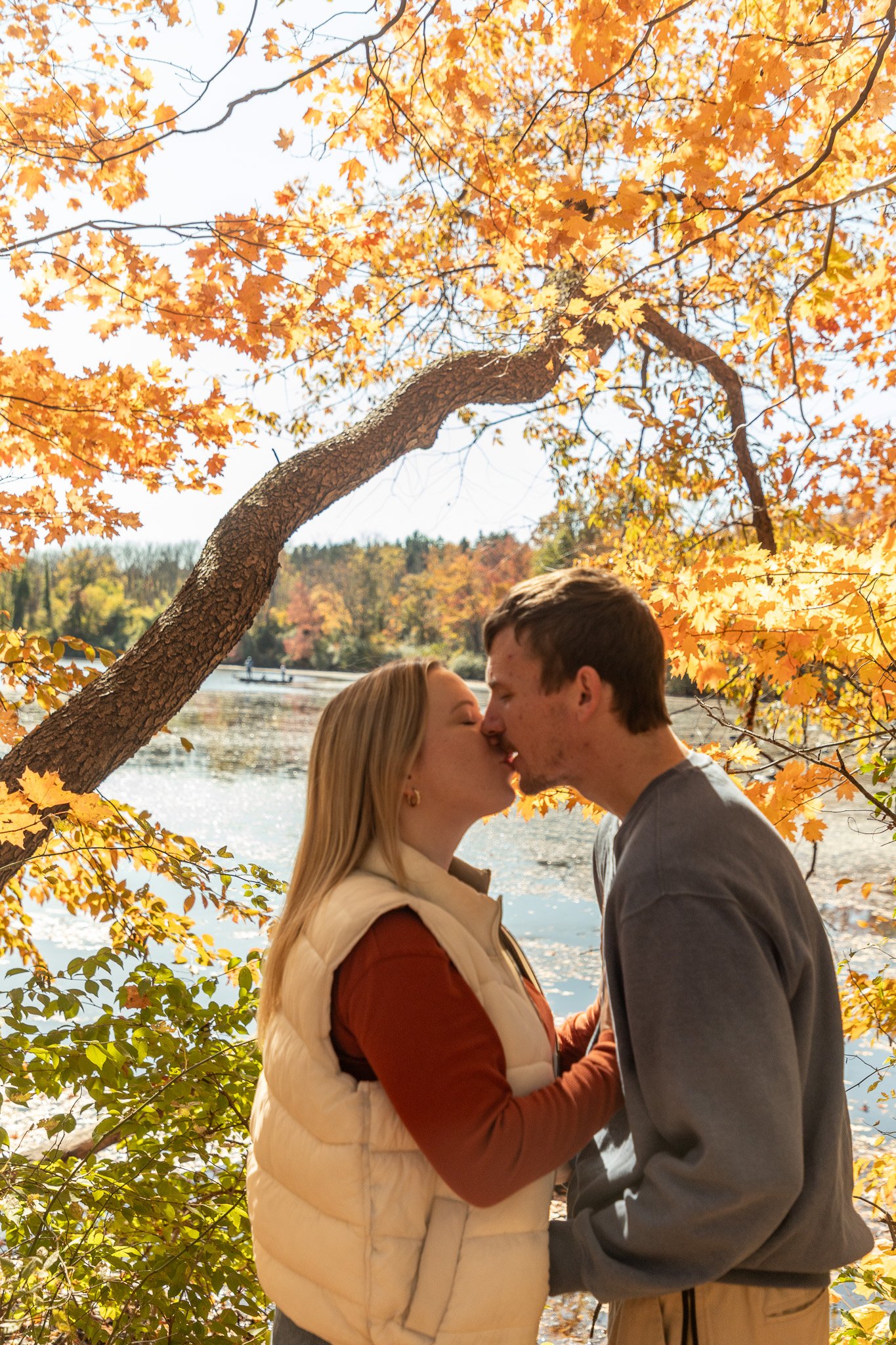 A young couple kissing outdoors near a lake during fall, surrounded by orange and yellow autumn leaves.