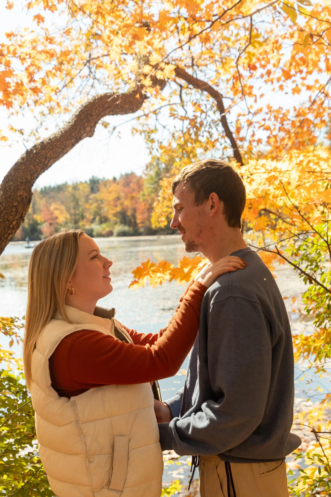 A couple stands close together outdoors near a lake in autumn, with fall foliage and a large leaning tree in the background. The woman has blonde hair and is wearing a beige vest and rust-colored sweatshirt. The man has brown hair and is wearing a gr