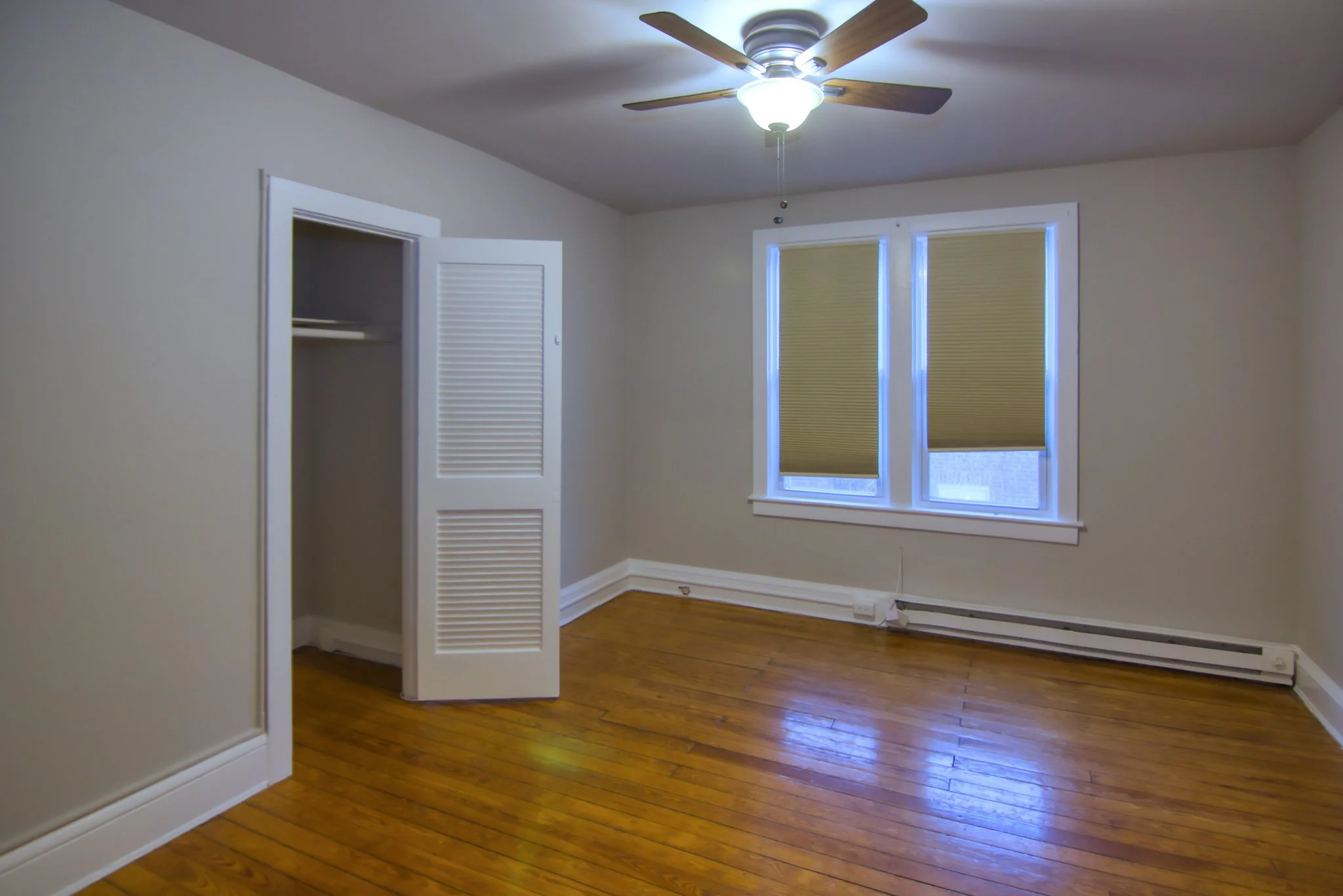Empty bedroom with hardwood floors, beige walls, a closet with louvered doors, two windows with brown blinds, a ceiling fan with lights, and baseboard heating.