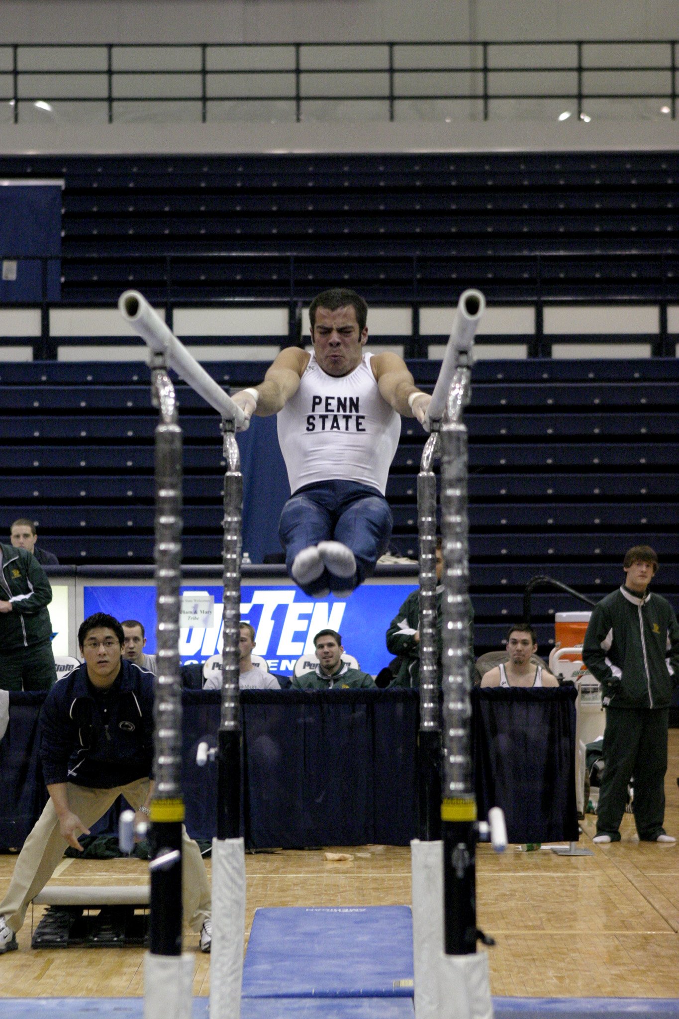 A Penn State male gymnast in a white tank top executing a routine on the parallel bars during a gymnastic competition, with coaches and spectators in the background.