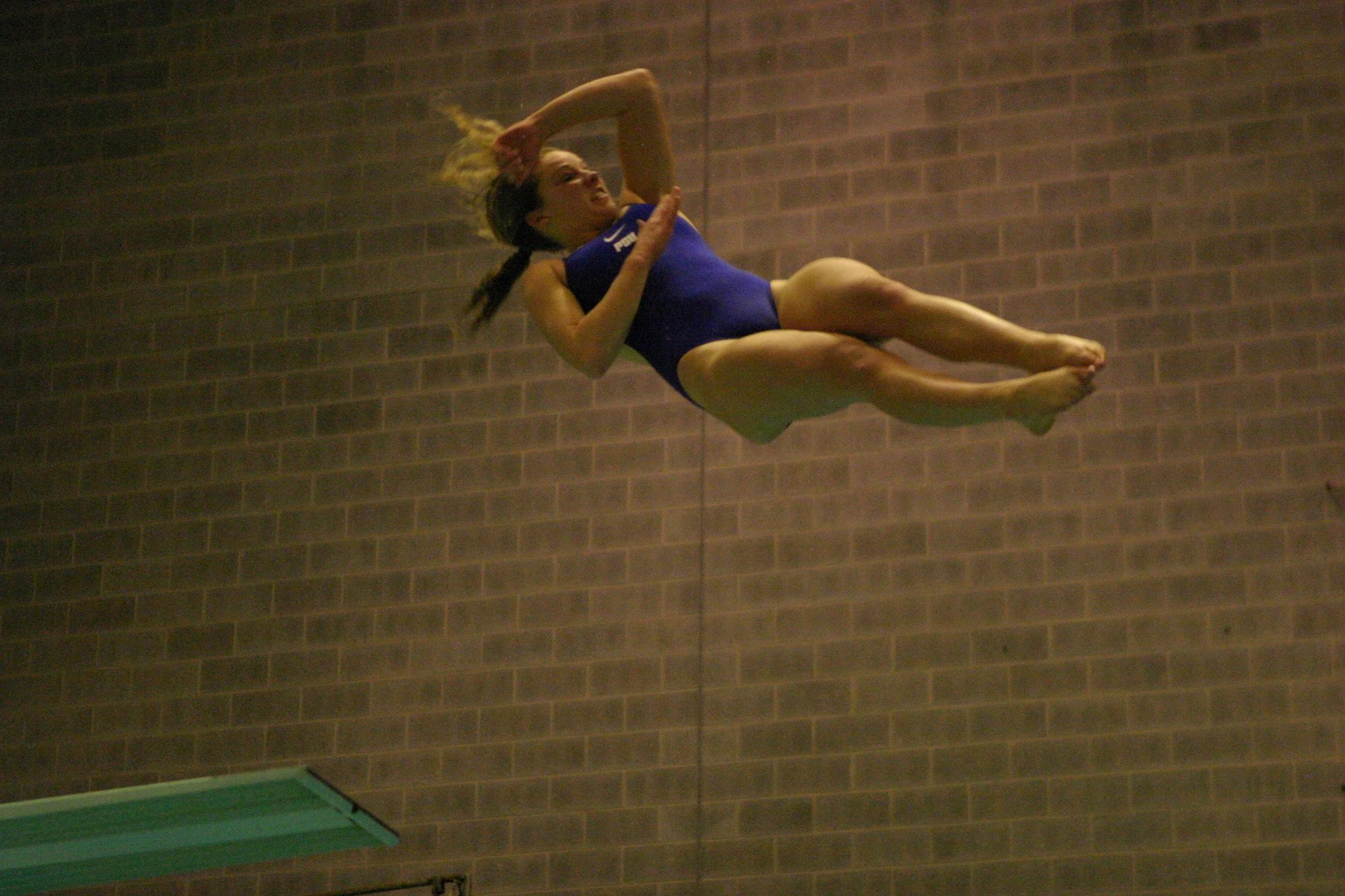A female gymnast in a blue leotard performs a flip in mid-air against a brick wall background.