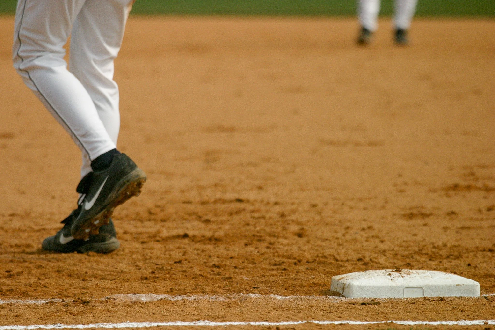 Close-up of a baseball player’s legs and feet on a dirt field near a base, with a player in the background.