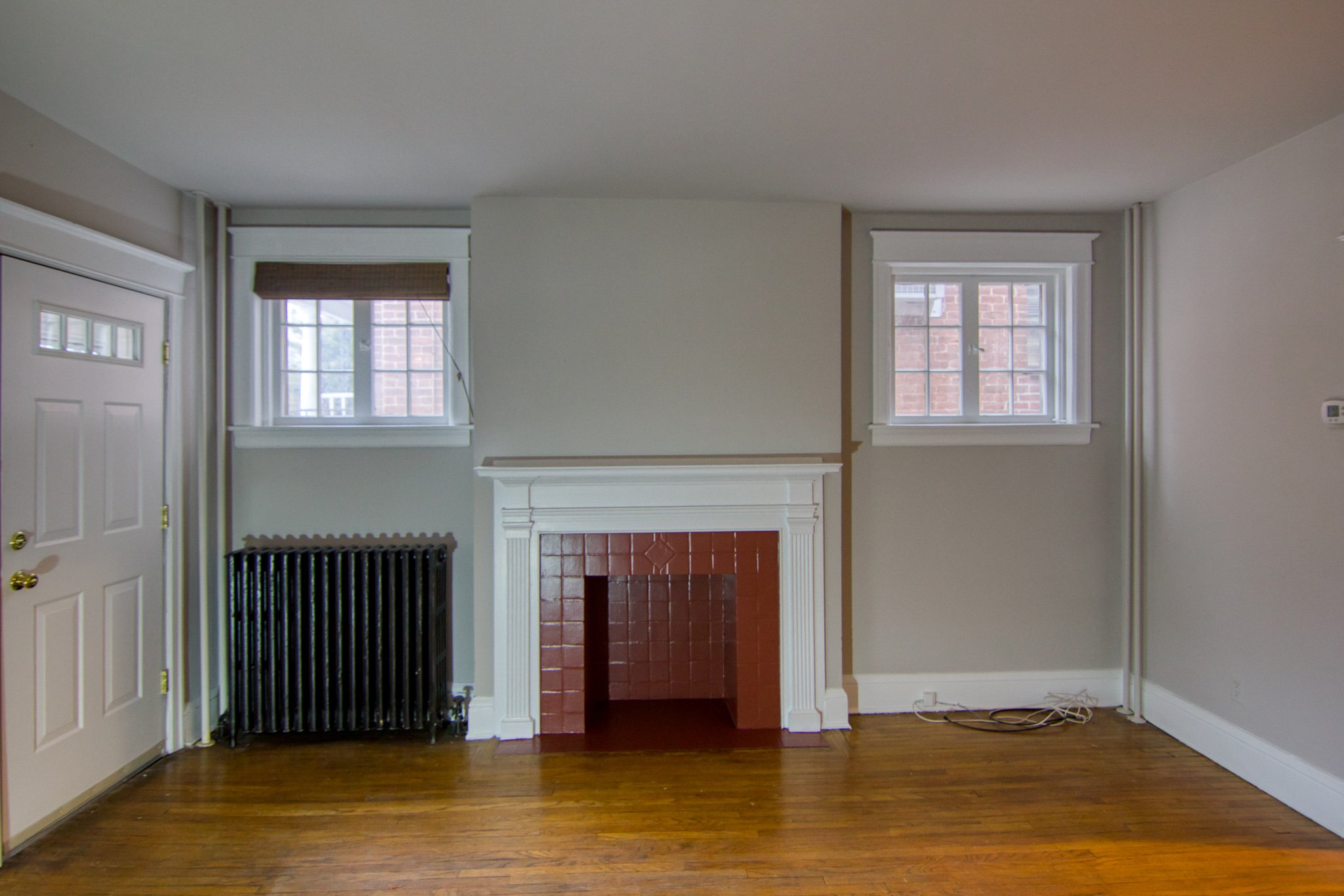 Empty living room with hardwood floors, a white fireplace with brick interior, two windows with blinds, radiator beneath one window, a closed door, and some electrical cords on the floor.