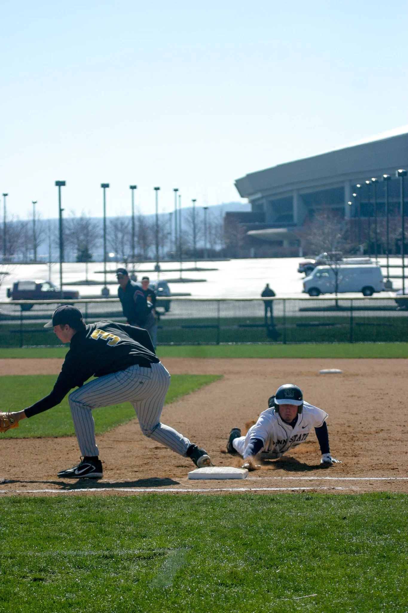 A baseball game with a player in a white uniform sliding into a base while another player in a black uniform tries to catch the ball. Two spectators and a few other players are visible in the background on a sunny day.