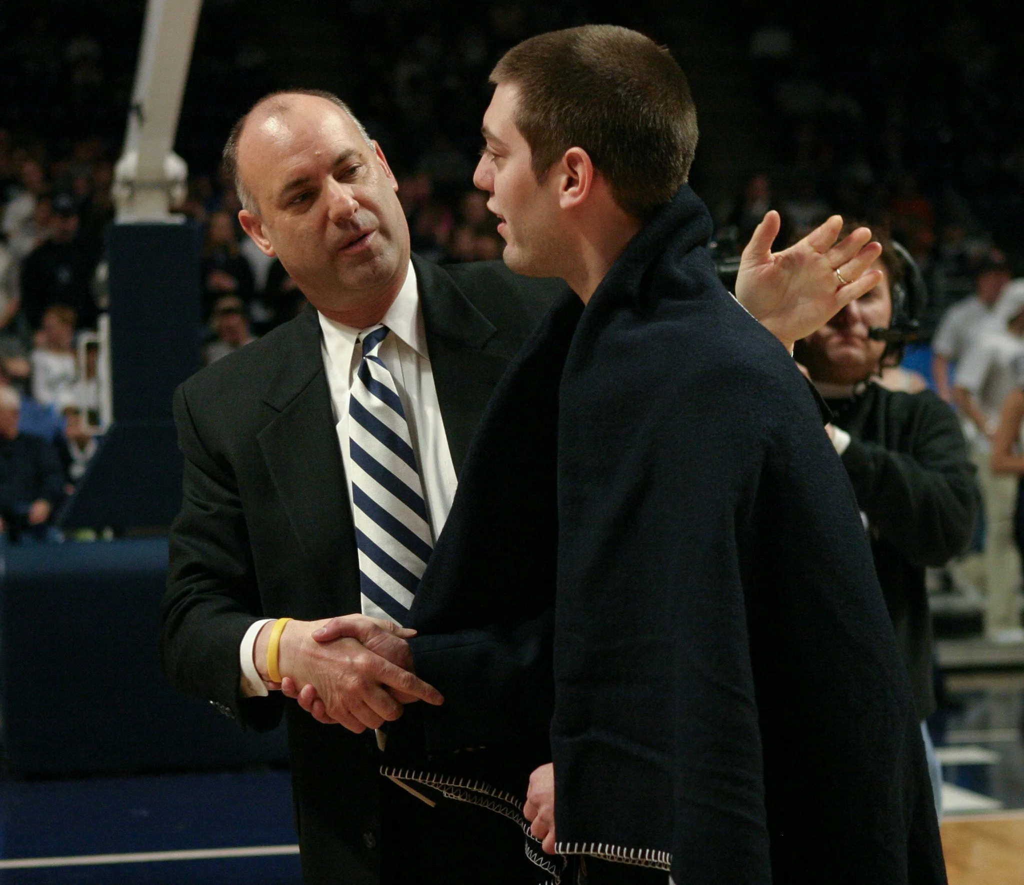 Two men, one in a black suit and striped tie, the other in a dark coat, shaking hands in a crowded indoor event space with a basketball court in the background.