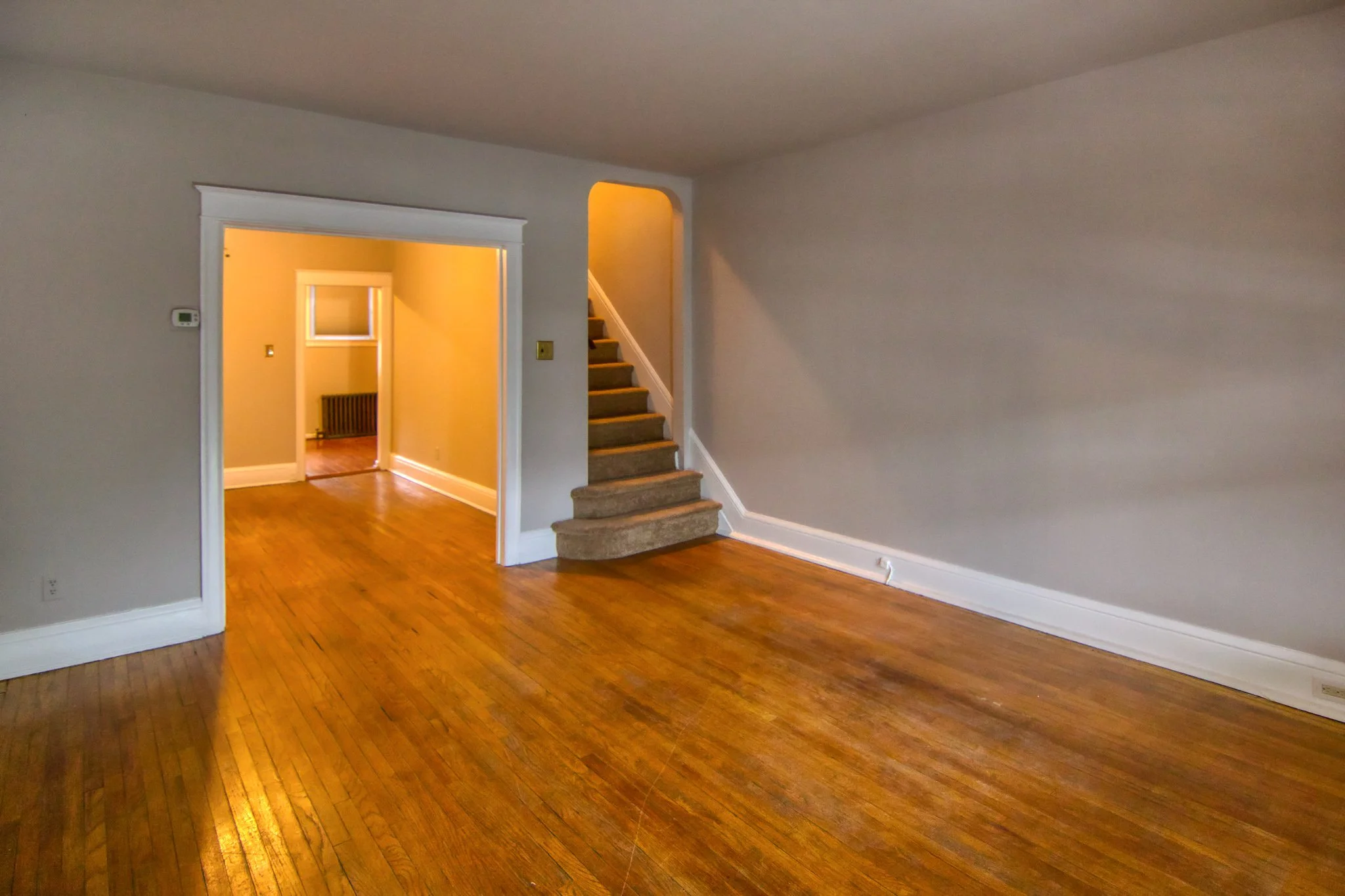 Empty living room with hardwood floors, gray walls, a staircase, and a doorway leading to another room with beige walls.