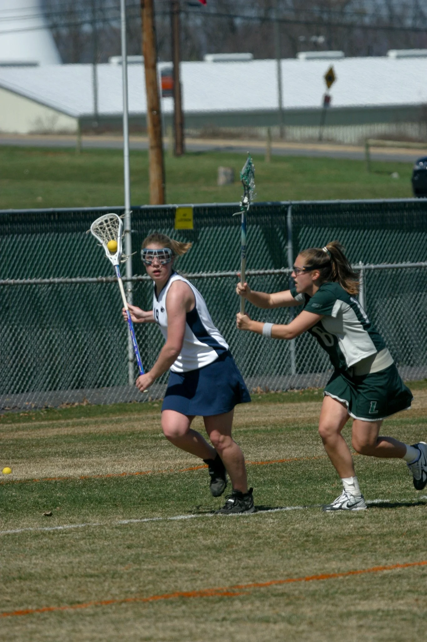 Two female lacrosse players competing for the ball on a grassy field with a fence and buildings in the background.