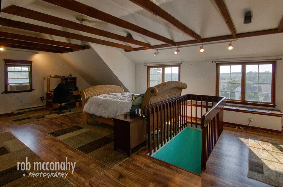 Living space with wooden floors, sloped ceiling with exposed beams, two windows, a couch, small desk with computer, and a stairwell with blue flooring.