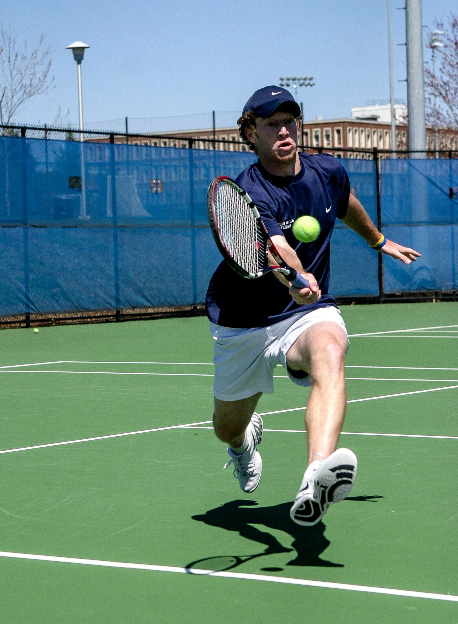 A male tennis player in a blue shirt, white shorts, and a black cap hitting a tennis ball with a racket on an outdoor court.
