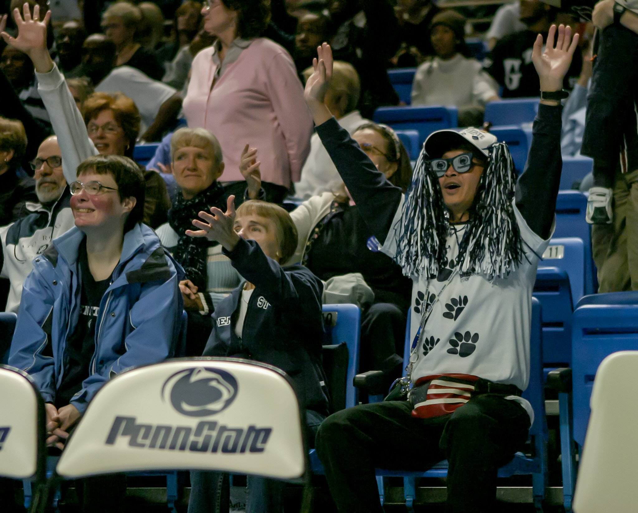 Group of people seated in a stadium with some raising their hands, one woman dressed as a fan with a wig, glasses, and a Penn State shirt cheering.