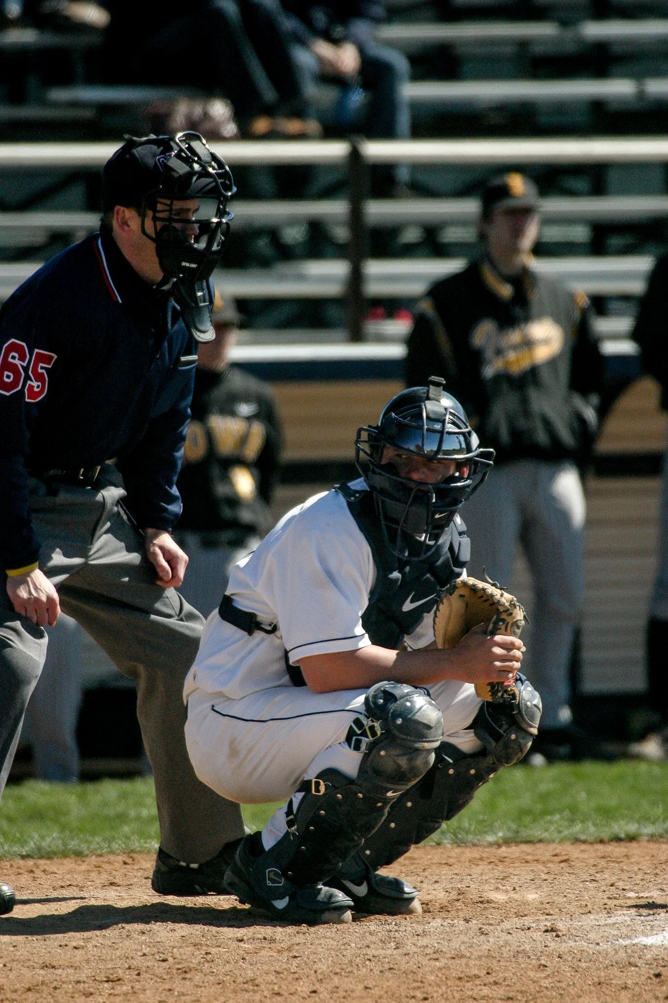 A baseball catcher in a crouched position behind the home plate, wearing a black helmet, black catcher's gear, and a white uniform with black accents. An umpire dressed in dark clothing stands next to the catcher, observing the game. The background i