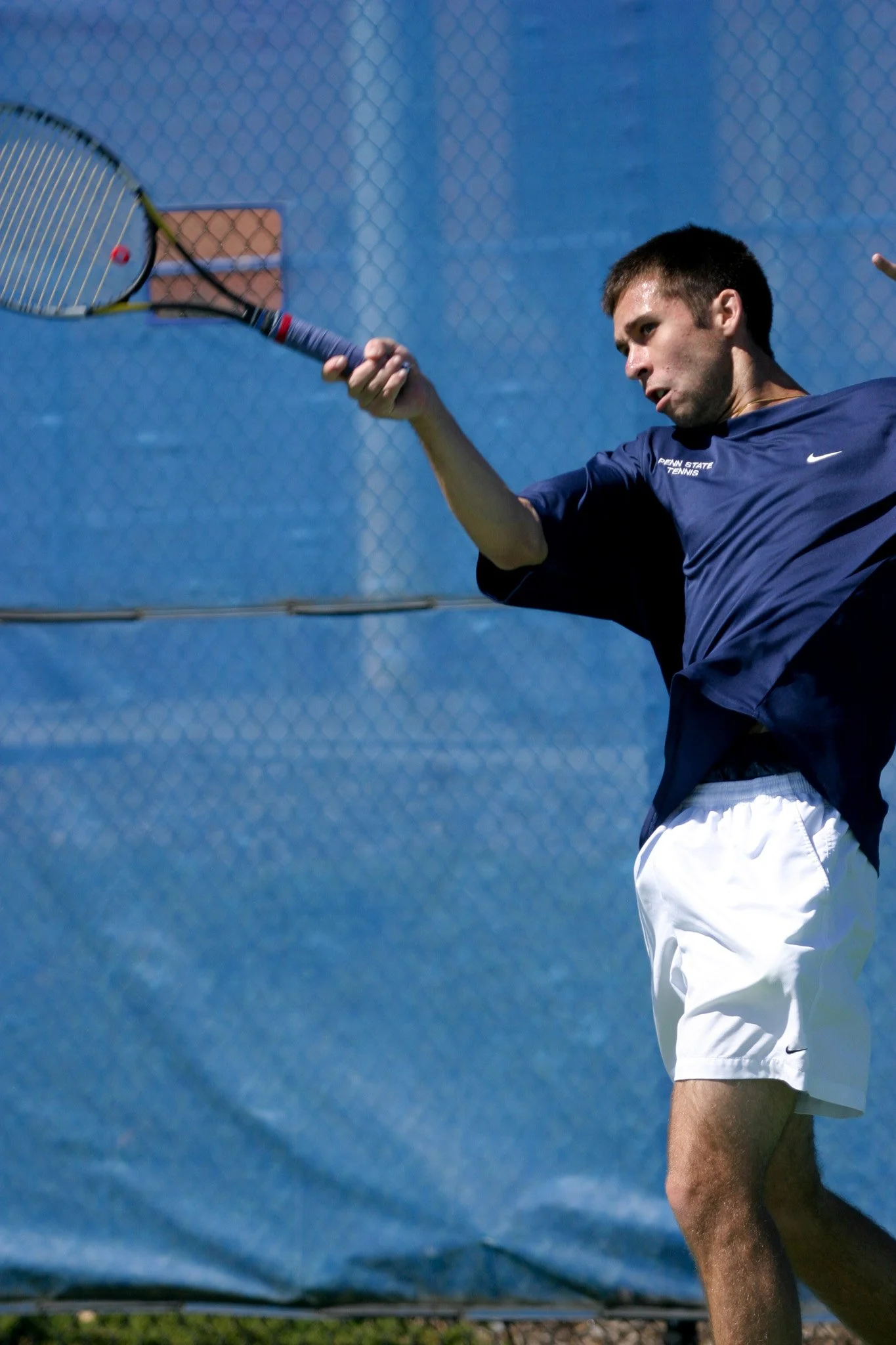 A man playing tennis on an outdoor court, wearing a navy blue shirt and white shorts, hitting a tennis ball with a racket.