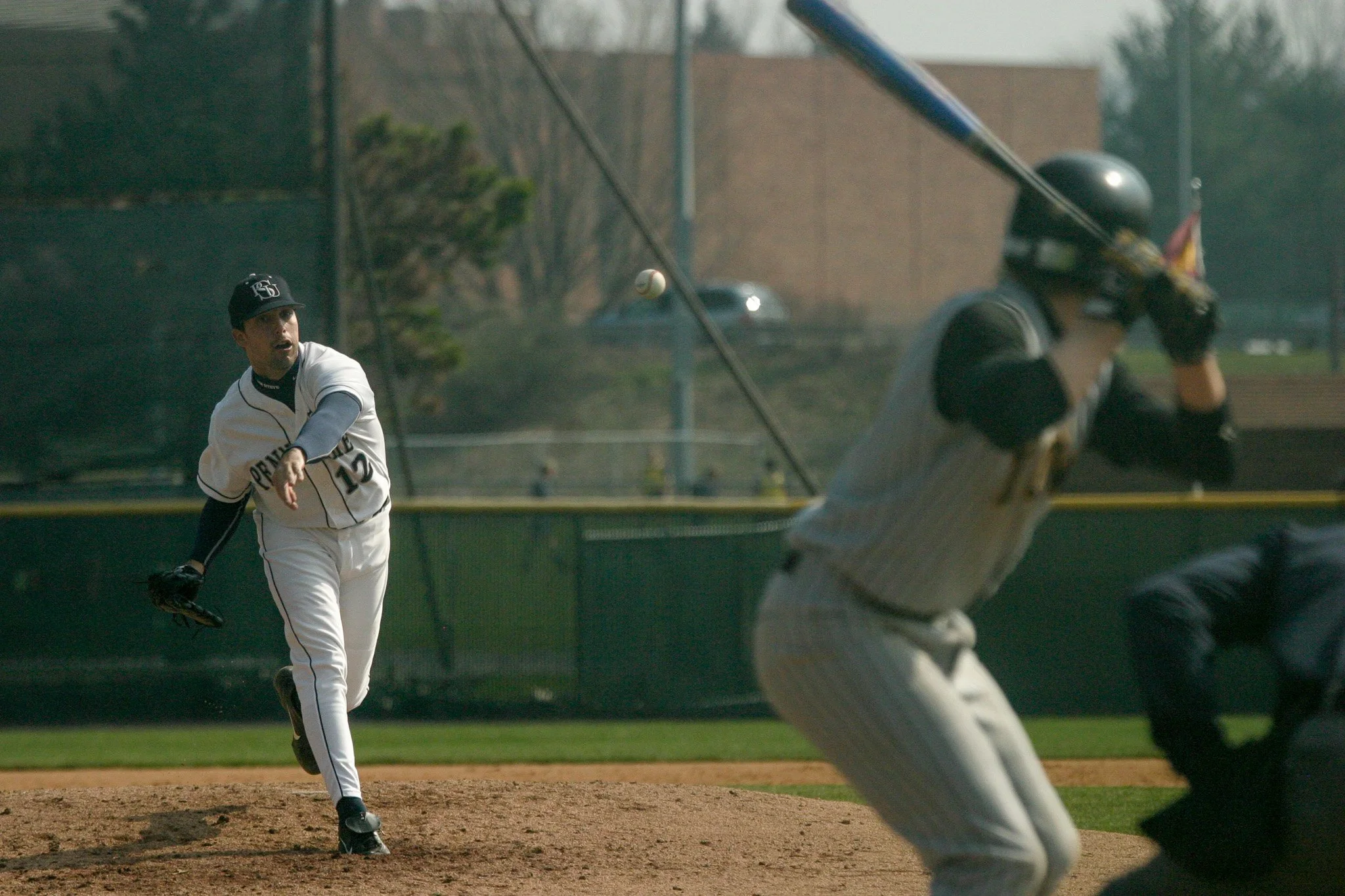 A baseball pitcher in a white uniform and cap throws a ball towards a batter standing at home plate. The batter is in a batting stance, wearing a gray pinstripe uniform and a helmet. An umpire crouches behind the batter, and the game is playing on a 