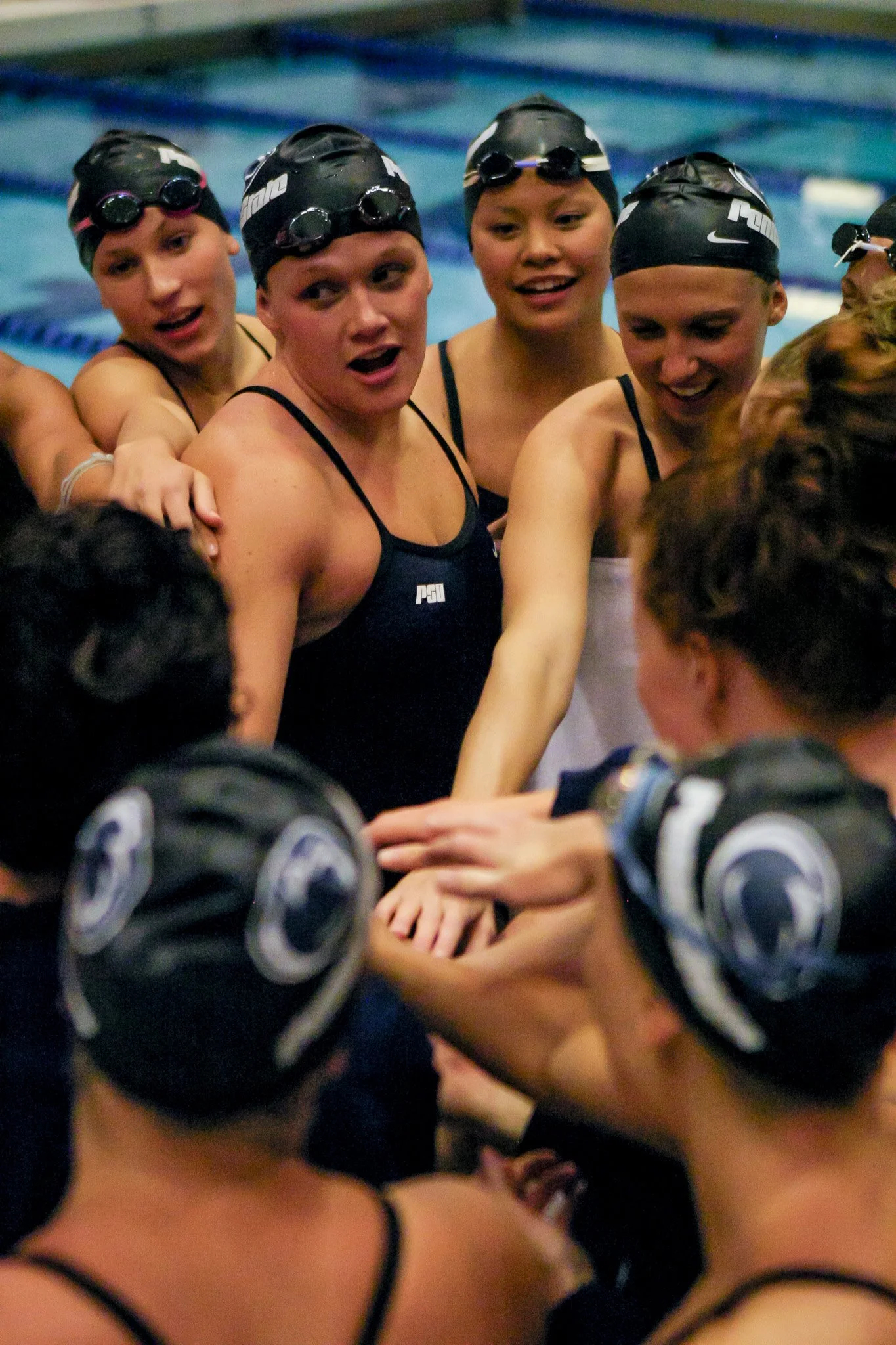 Swimmers in a huddle during a team meeting at a swimming pool, wearing swim caps and racing suits.