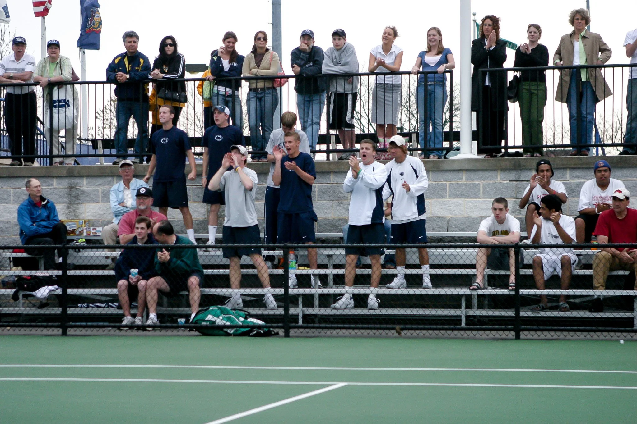 People watching a tennis match from bleachers, some standing and cheering, others sitting and clapping.