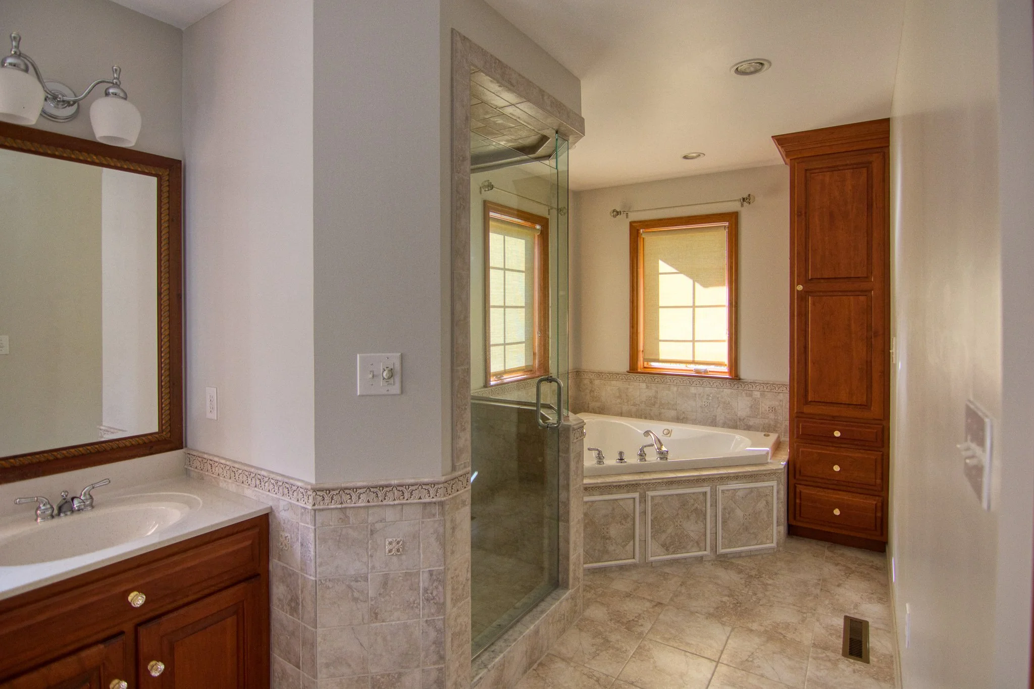 Bathroom with a single sink vanity, mirror, wall-mounted light, tiled tub surround, large corner jacuzzi tub, window, and glass-enclosed shower.