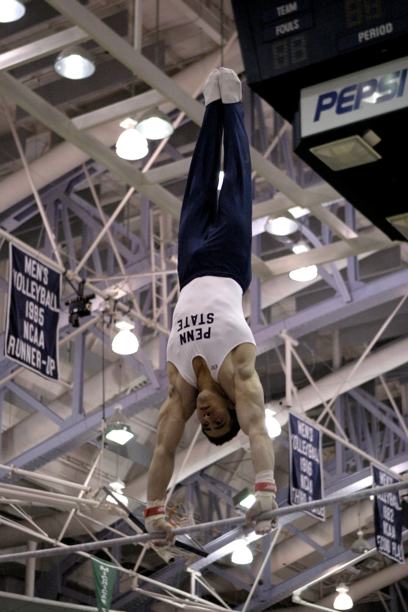 Male gymnast in a white tank top and dark pants performing a handstand on parallel bars in a gymnasium with banners hanging from the ceiling.
