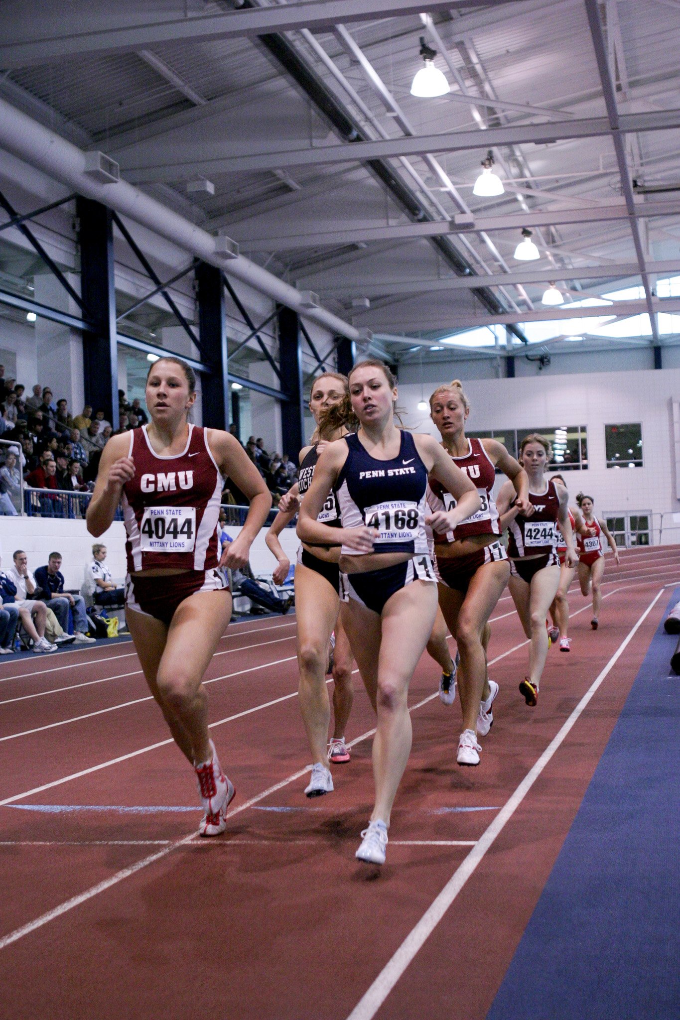 Group of female athletes competing in a track race inside an indoor stadium with spectators watching from the sidelines.