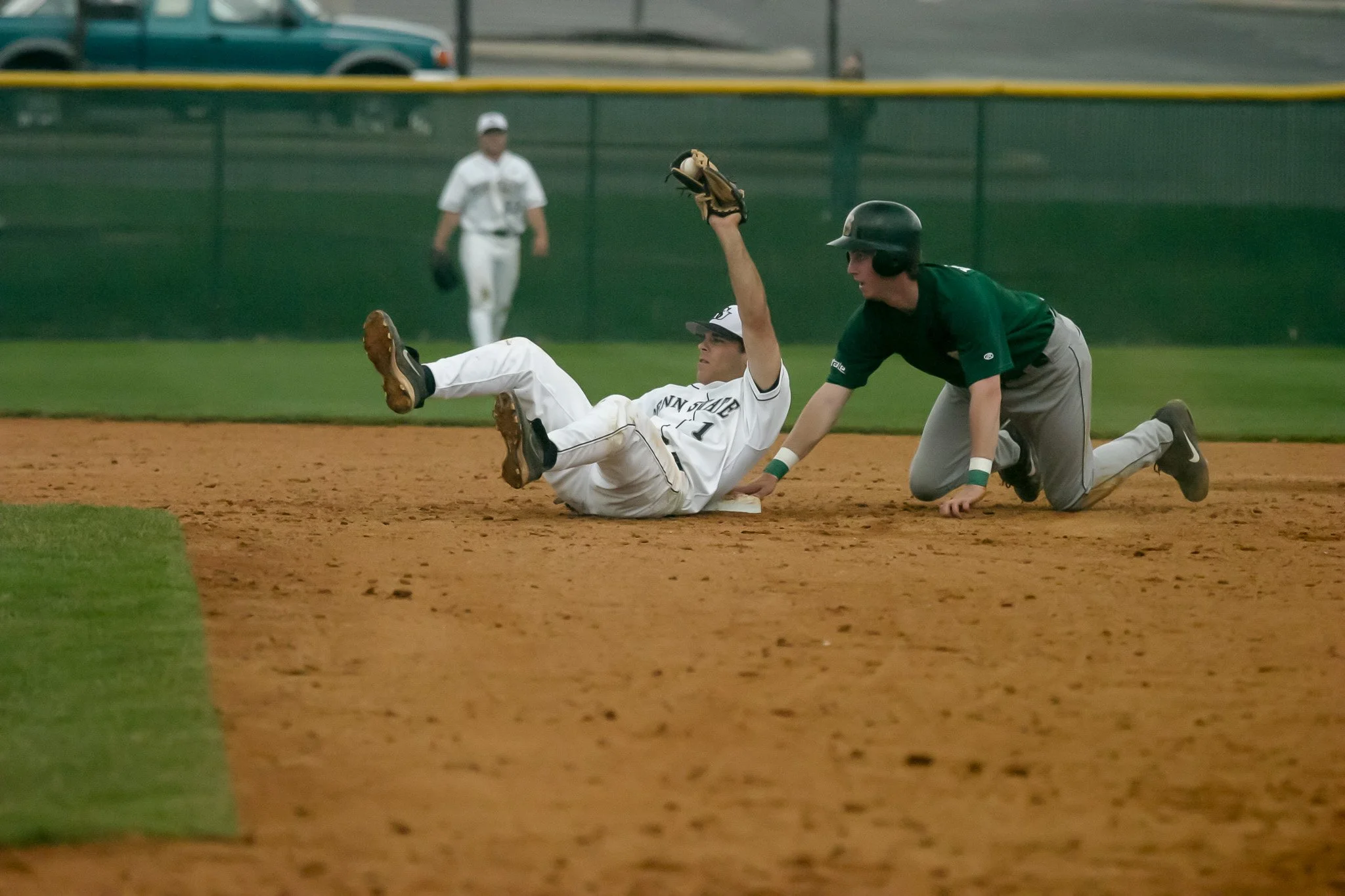 A baseball player in a white uniform lying on the ground after a play, with a baseball glove, while another player in a green shirt and gray pants is kneeling nearby on the dirt field.
