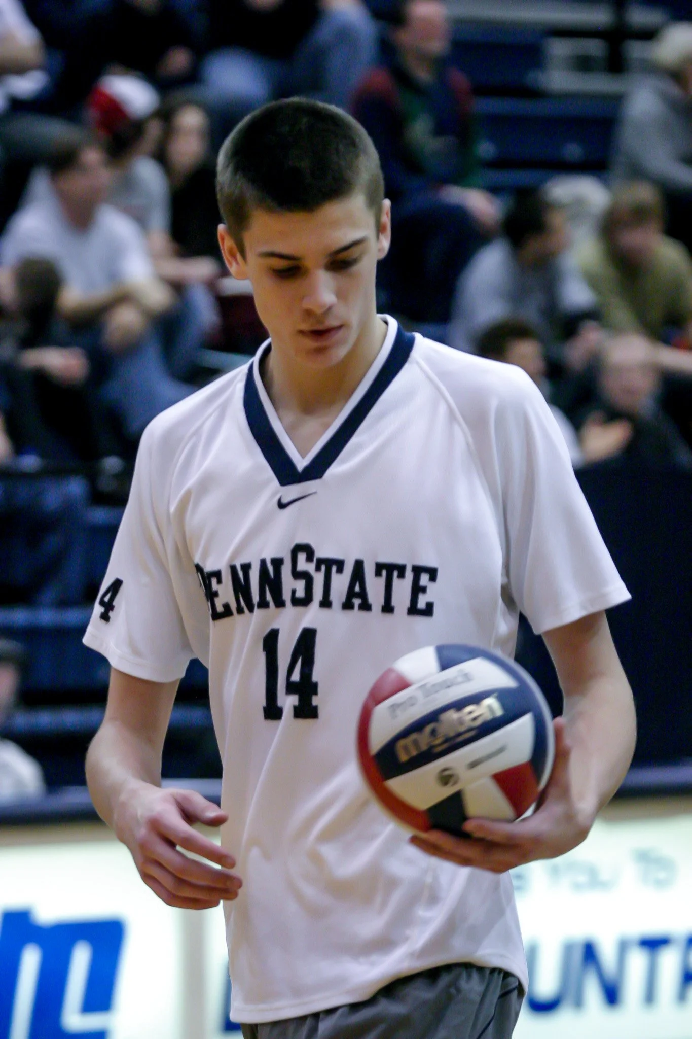 Young man in a Penn State basketball jersey holding a volleyball during a game, with spectators in the background.