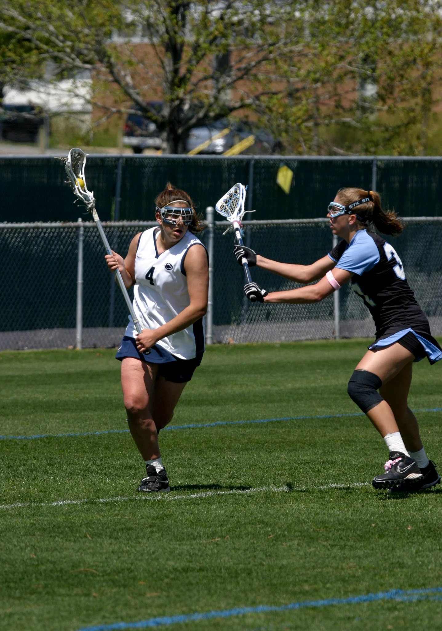 Two women playing lacrosse on a grassy field, one in a white jersey and the other in a black and blue jersey. The woman in white appears to be avoiding a check from the woman in black and blue.