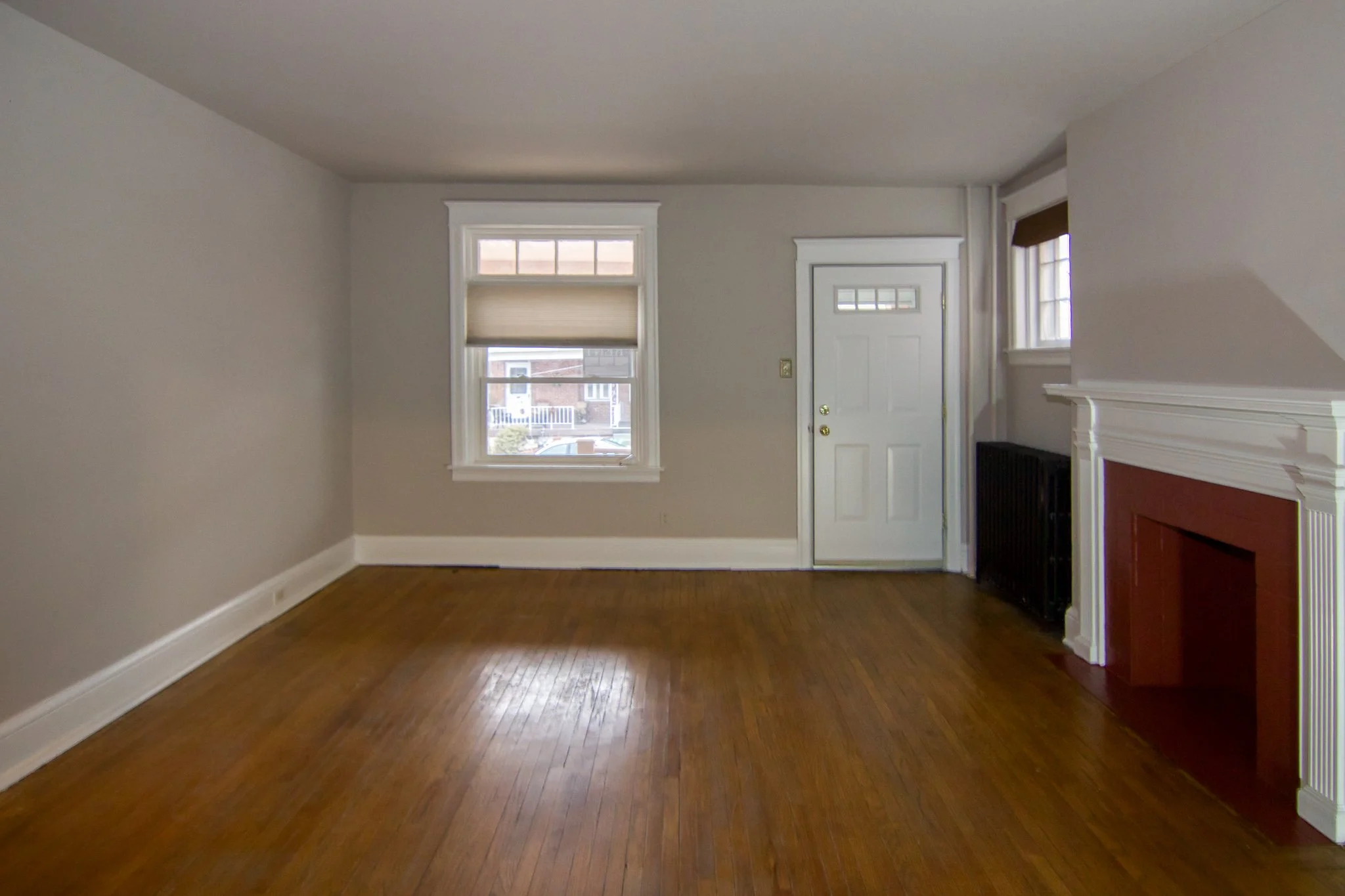 Empty living room with hardwood floors, two windows, a white front door, and a fireplace with a red hearth.