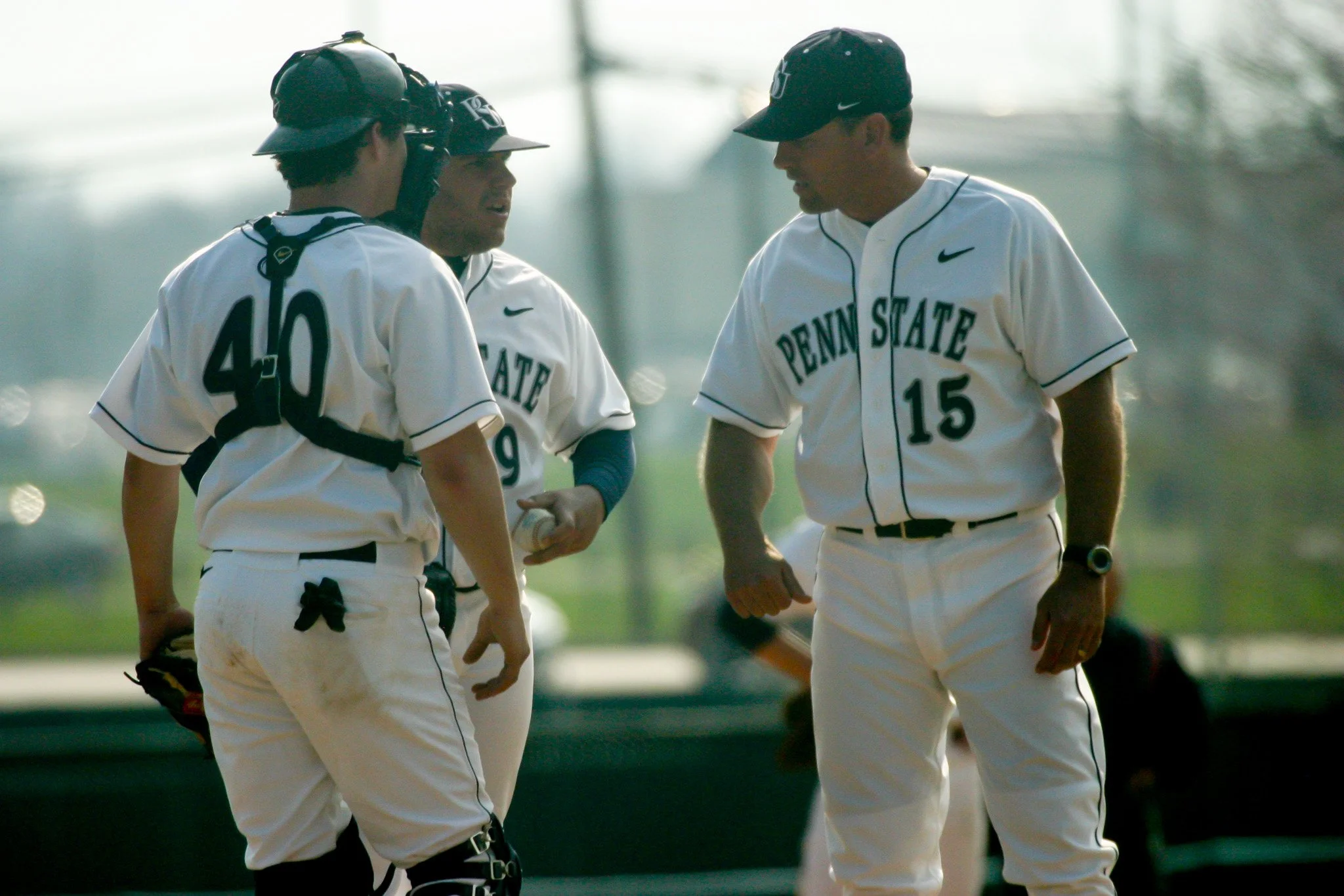 Four baseball players from Penn State team in white uniforms, standing in a huddle.