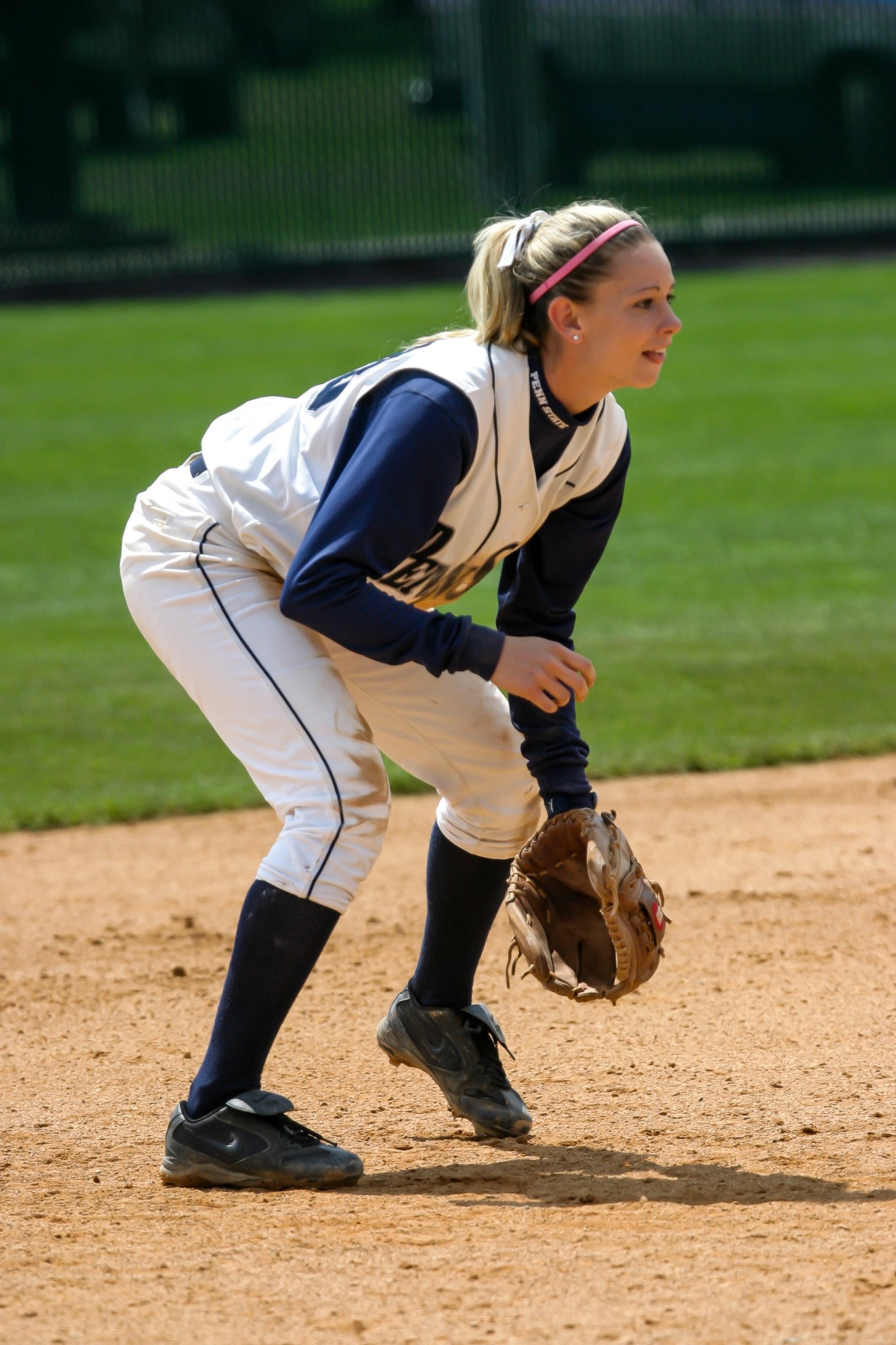 A female softball player wearing a white and navy uniform, with a pink headband, is in a ready position on the field, holding a glove.