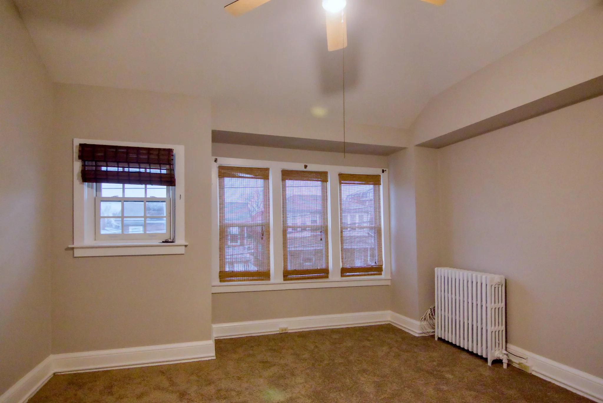 Empty room with beige walls, brown carpet, three large windows with bamboo shades, one smaller window with a brown blind, a ceiling fan with attached light, a white radiator, and some electrical cords on the floor.