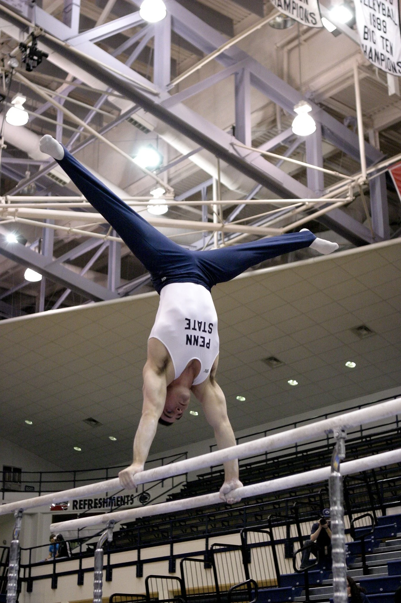 A male gymnast performing a handstand on the parallel bars in an indoor gymnasium, wearing a white tank top with "PENN STATE" printed on it and navy blue pants.