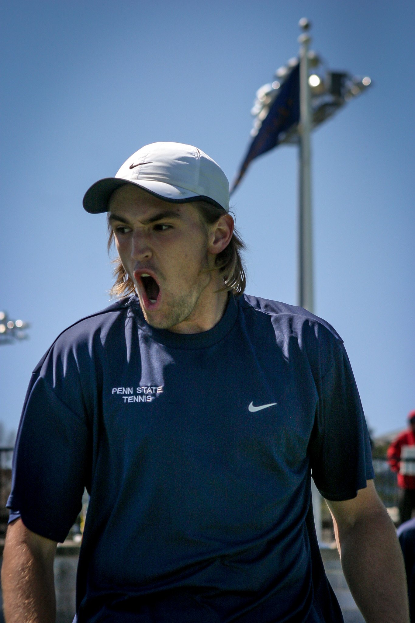 A young man wearing a white cap and a navy blue Penn State tennis shirt is shouting or cheering outdoors on a sunny day, with flags and stadium lights in the background.