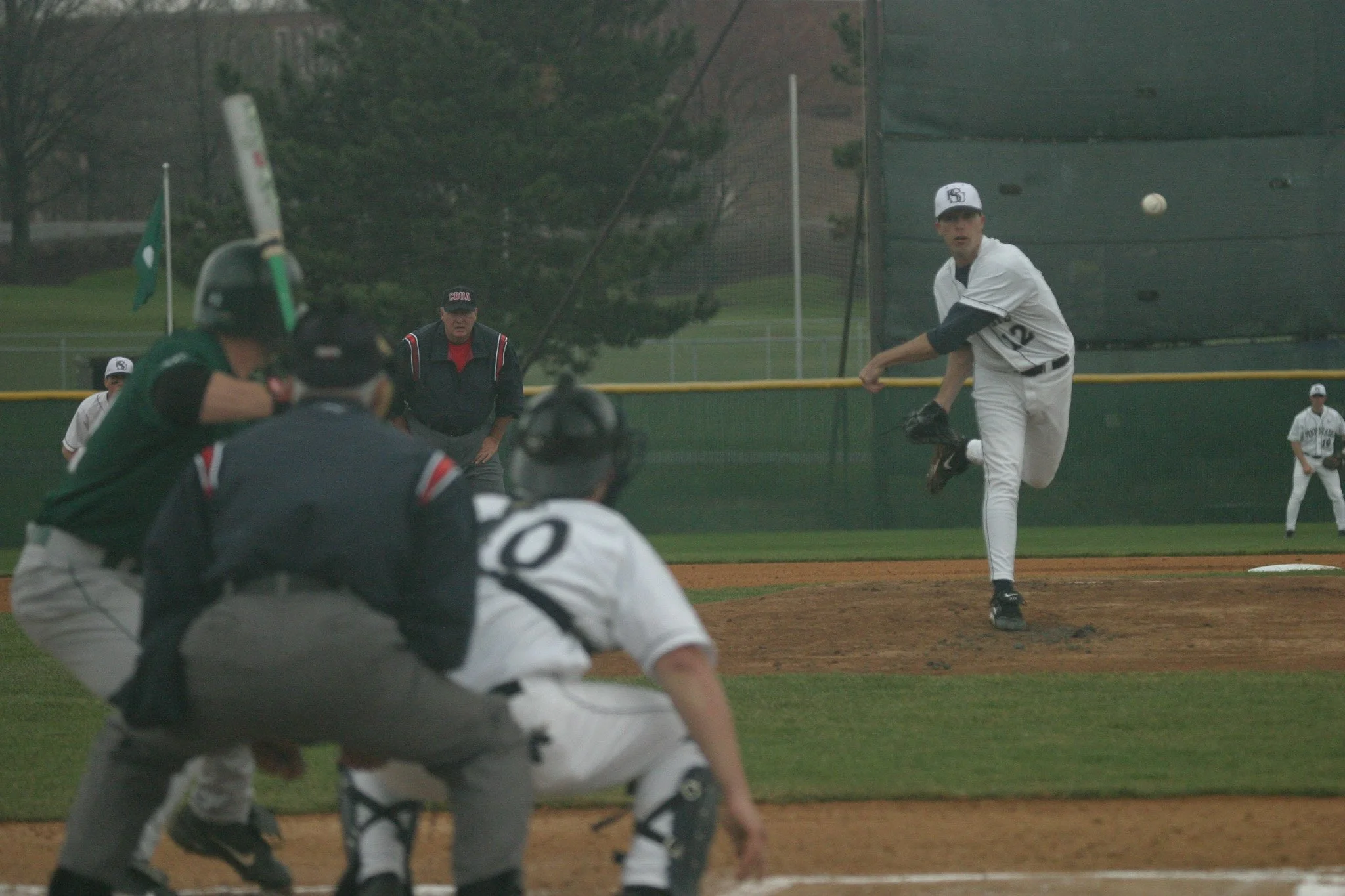 A baseball player in a white uniform with the number 12 is pitching the ball on a baseball field, with the batter and umpire also visible, and an outfield fence in the background.