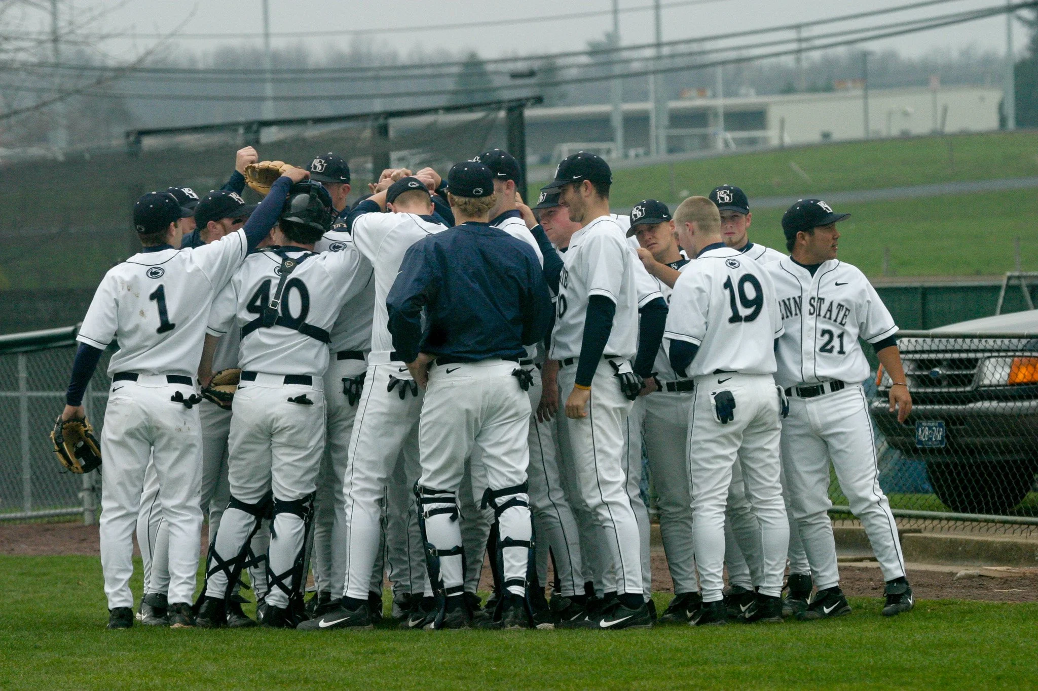 A group of baseball players in white uniforms with black caps gathered in a huddle on the field during daytime.