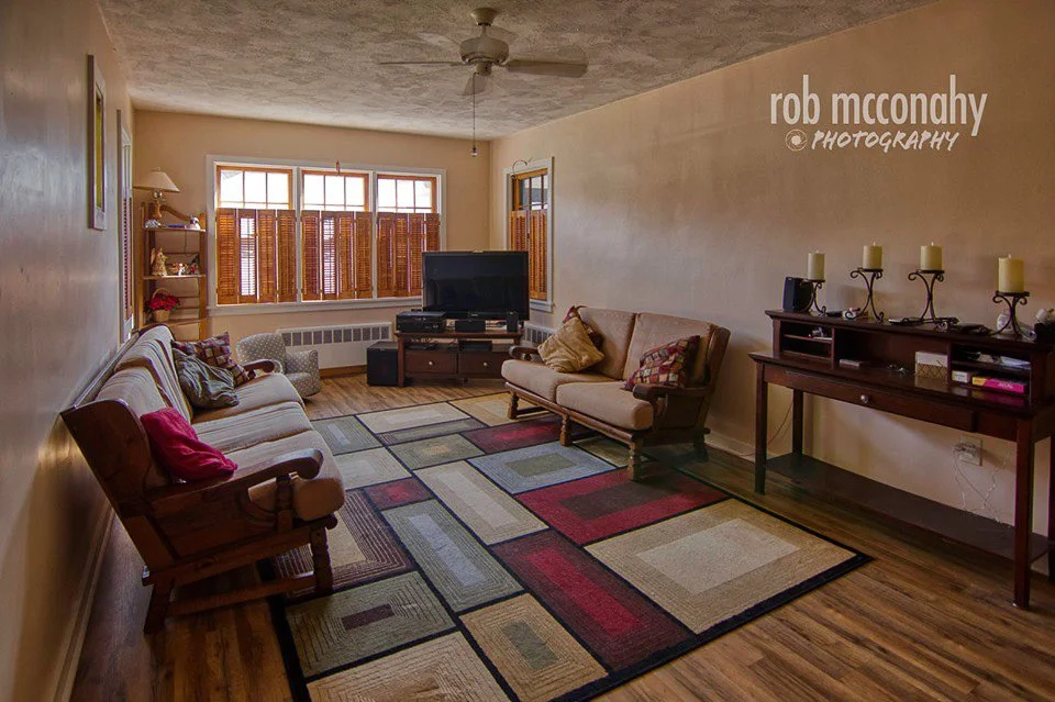 Living room with beige walls, hardwood floor, patterned area rug, three sofas, a TV on a stand, and a wooden console table with candles.