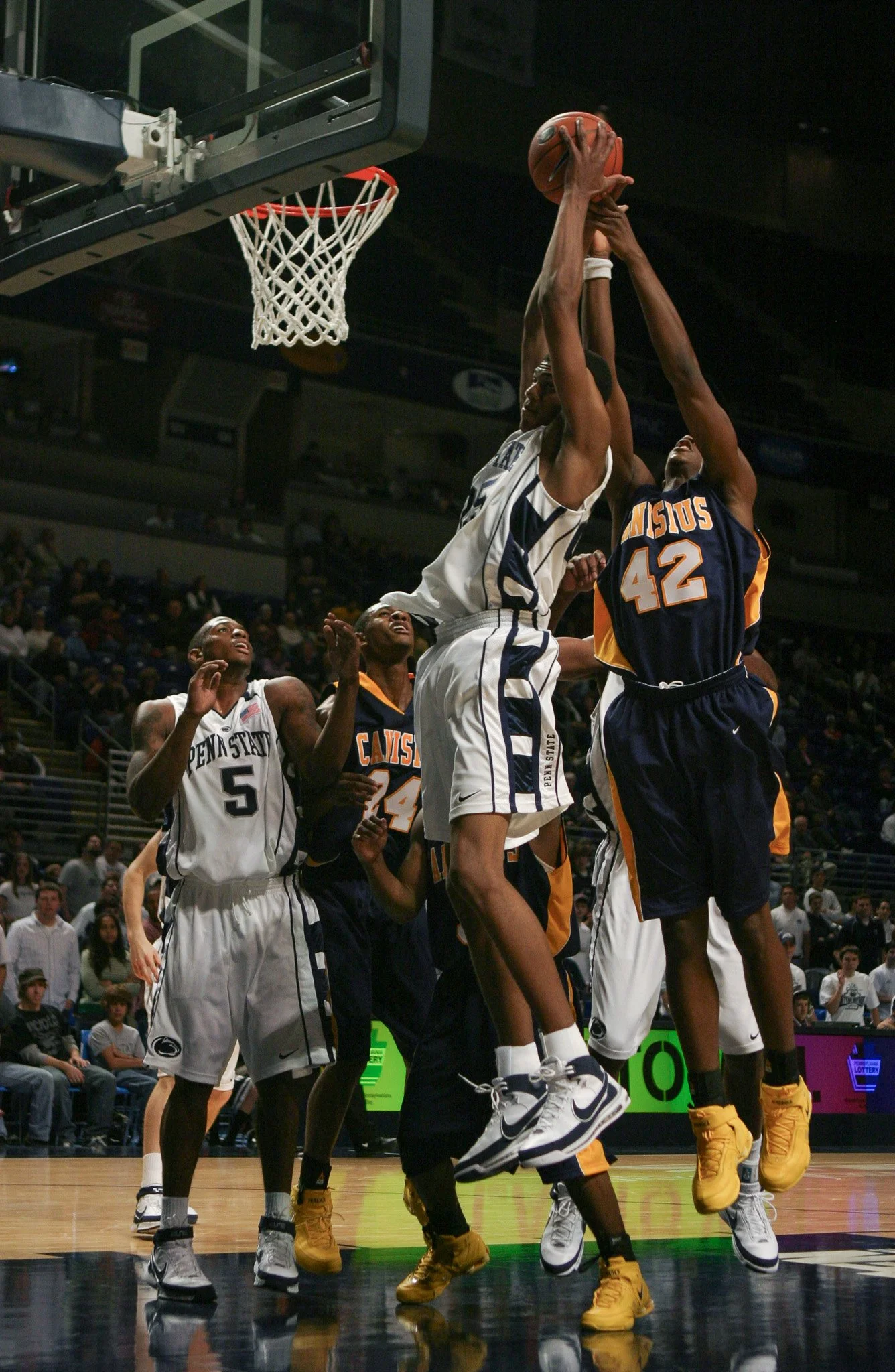 Basketball game with players jumping near the hoop, one player in white attempting a shot or rebound, and players in dark uniforms defending.
