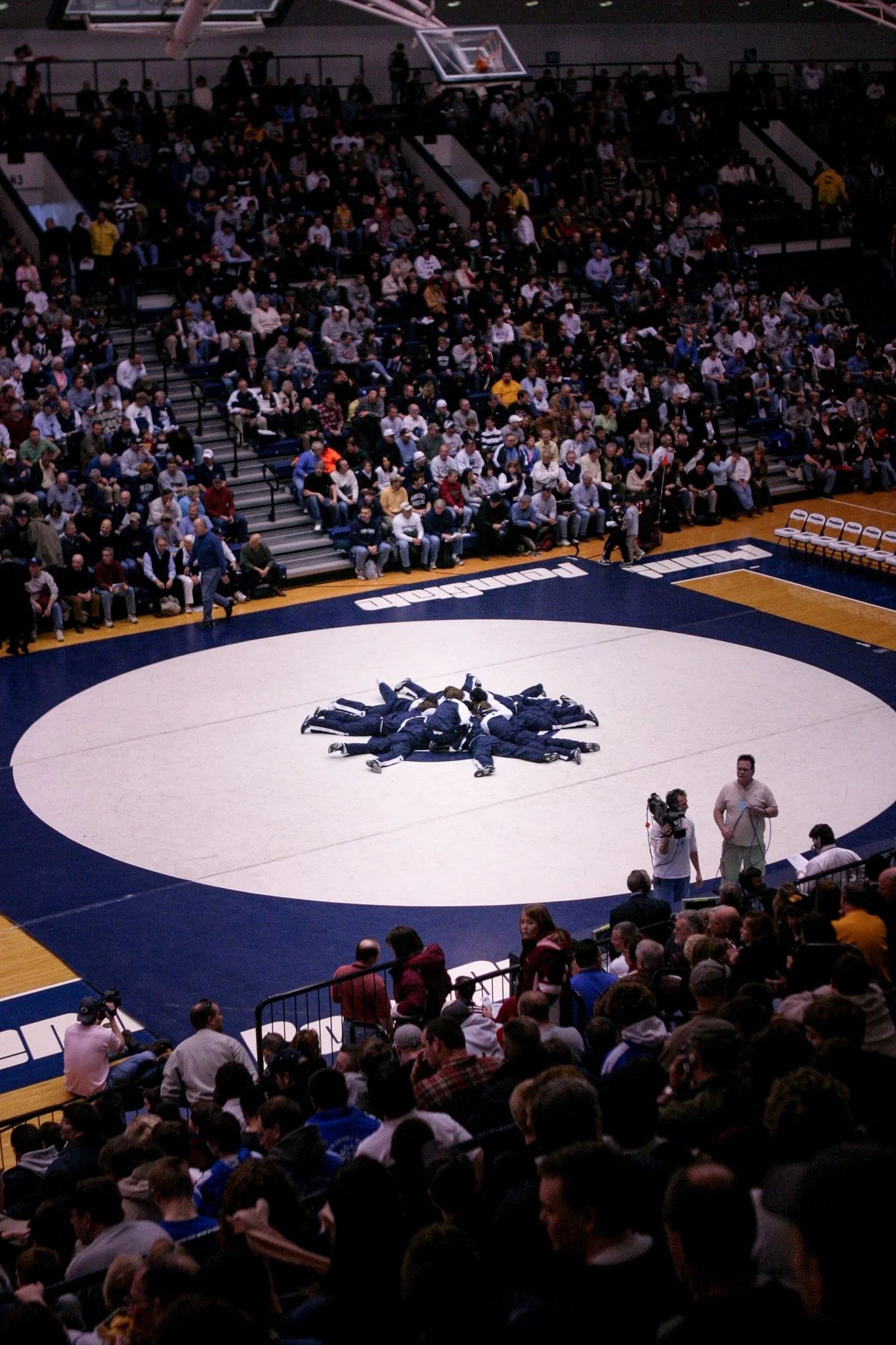 Wrestlers forming a circle on a wrestling mat during a match with a crowd watching in the stands.