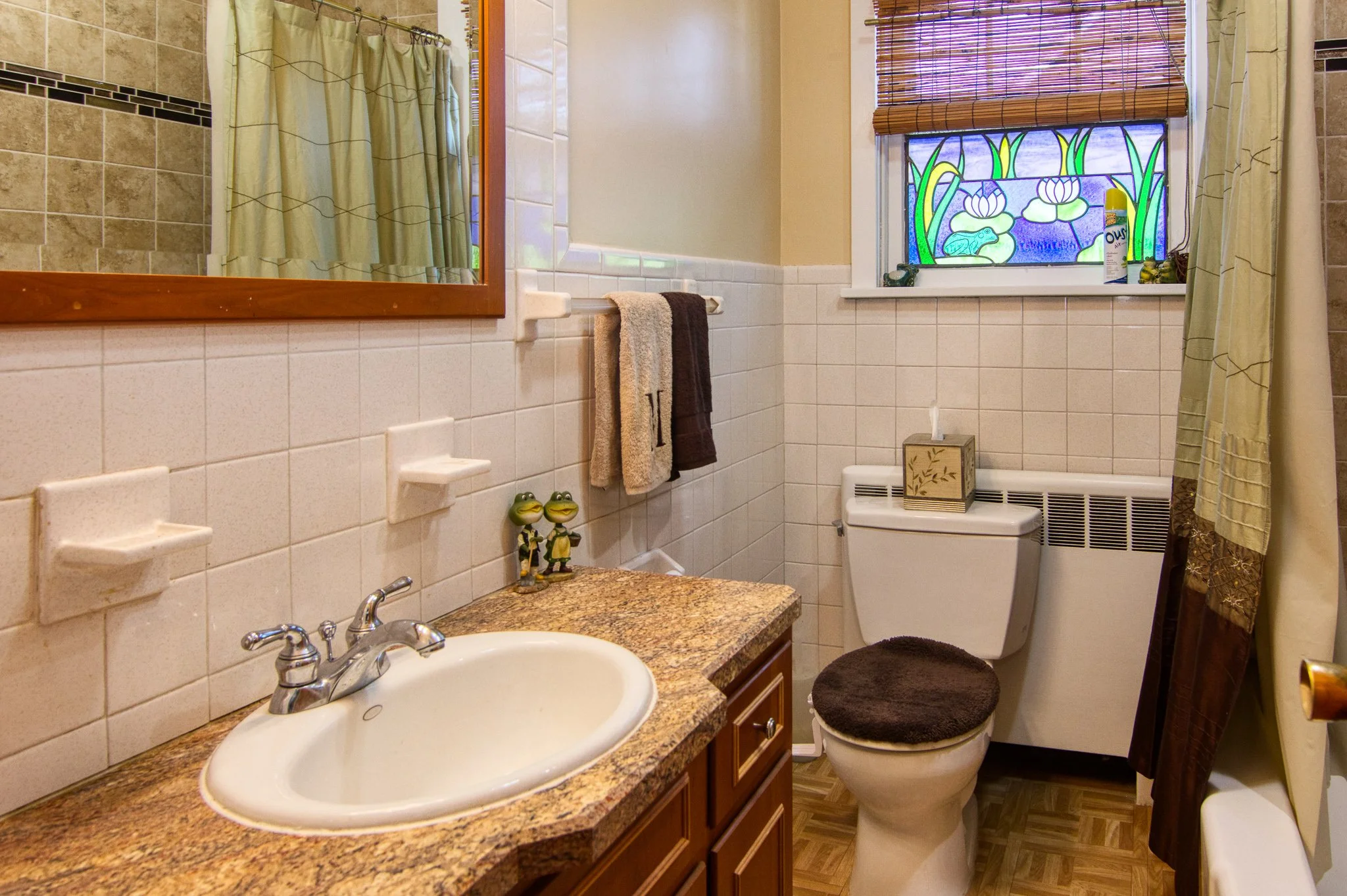 A small bathroom with a granite countertop sink, a large mirror, and a toilet. There are two frog figurines on the counter, a towel rack with brown and beige towels, a stained glass window, and a window shade. The bathroom has beige tiles with black 