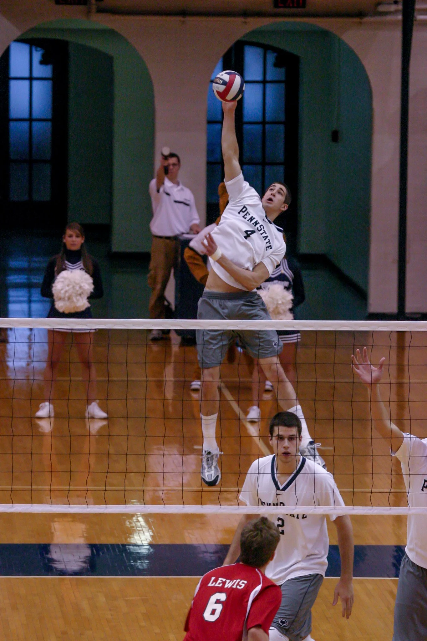 A volleyball player from Penn State is jumping to hit the ball over the net during a game, with other players and a cheerleader in the background.