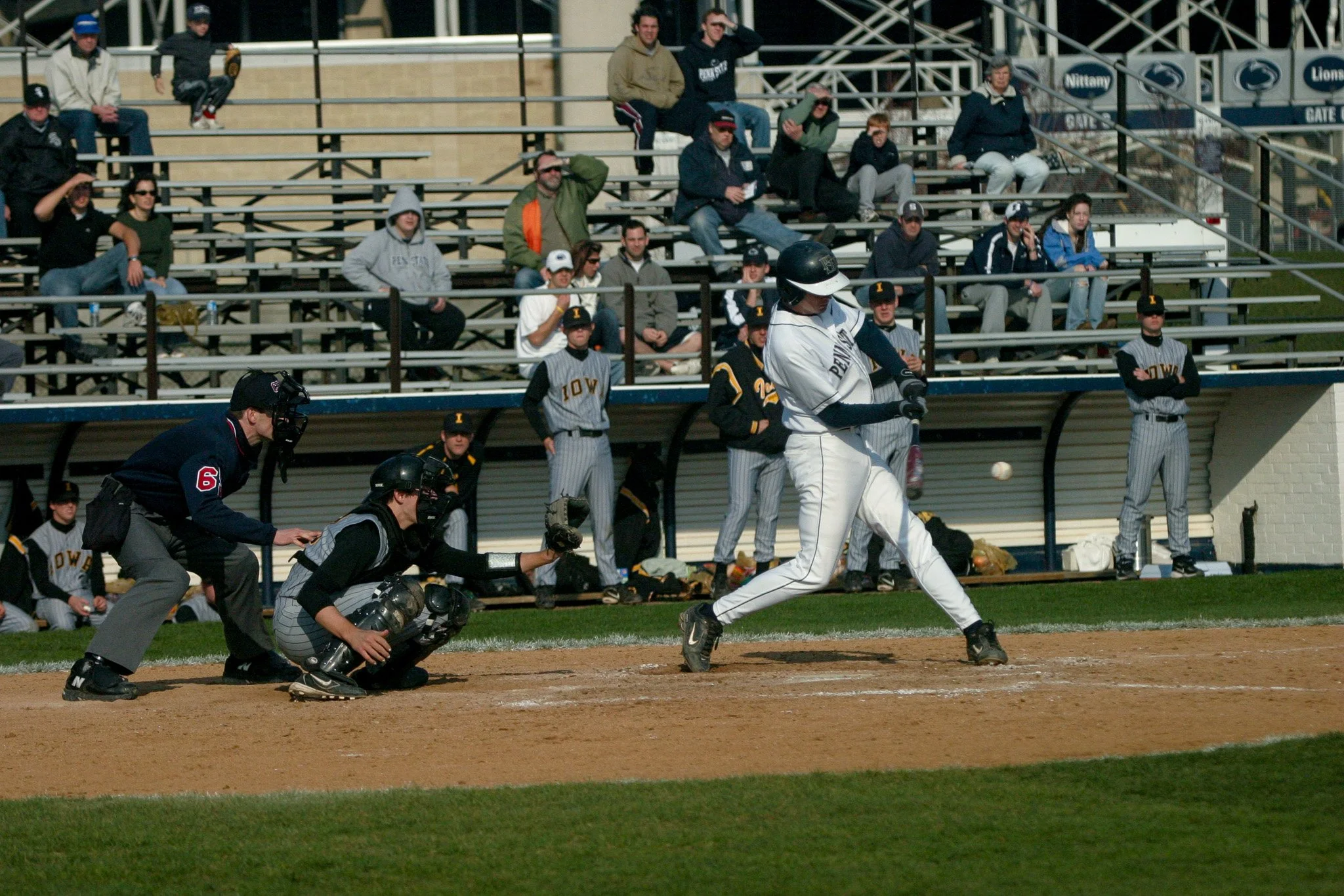A baseball player in a white uniform swings at a pitched ball, while a catcher and umpire crouch behind him. Spectators sit on bleachers in the background.