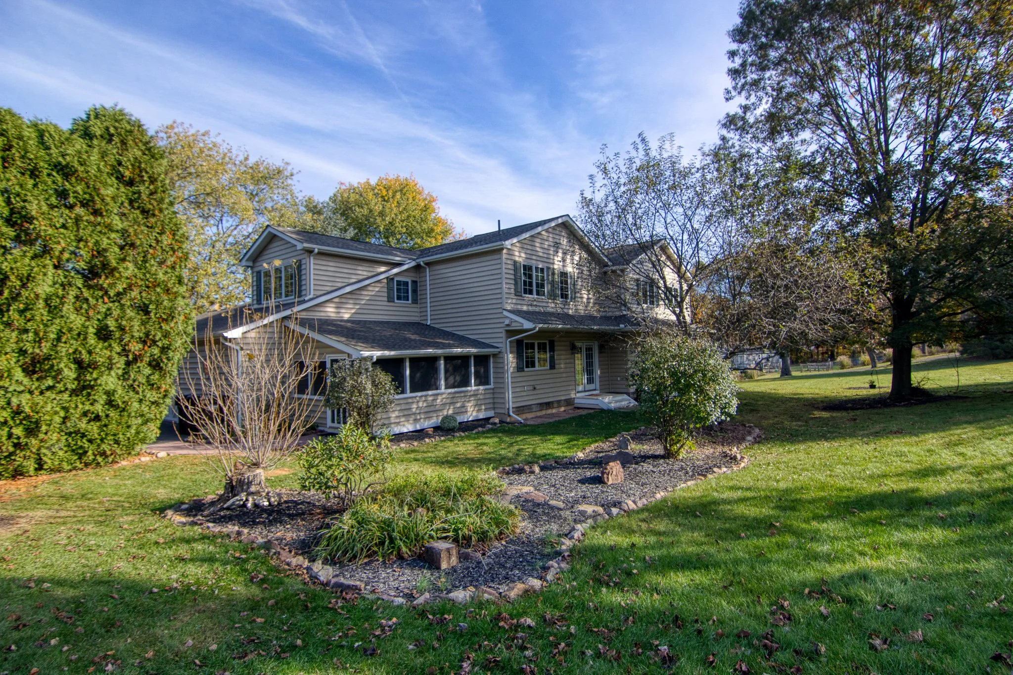 A two-story suburban house with beige siding and a dark roof, surrounded by a green lawn and trees with some leaves falling, under a partly cloudy blue sky.