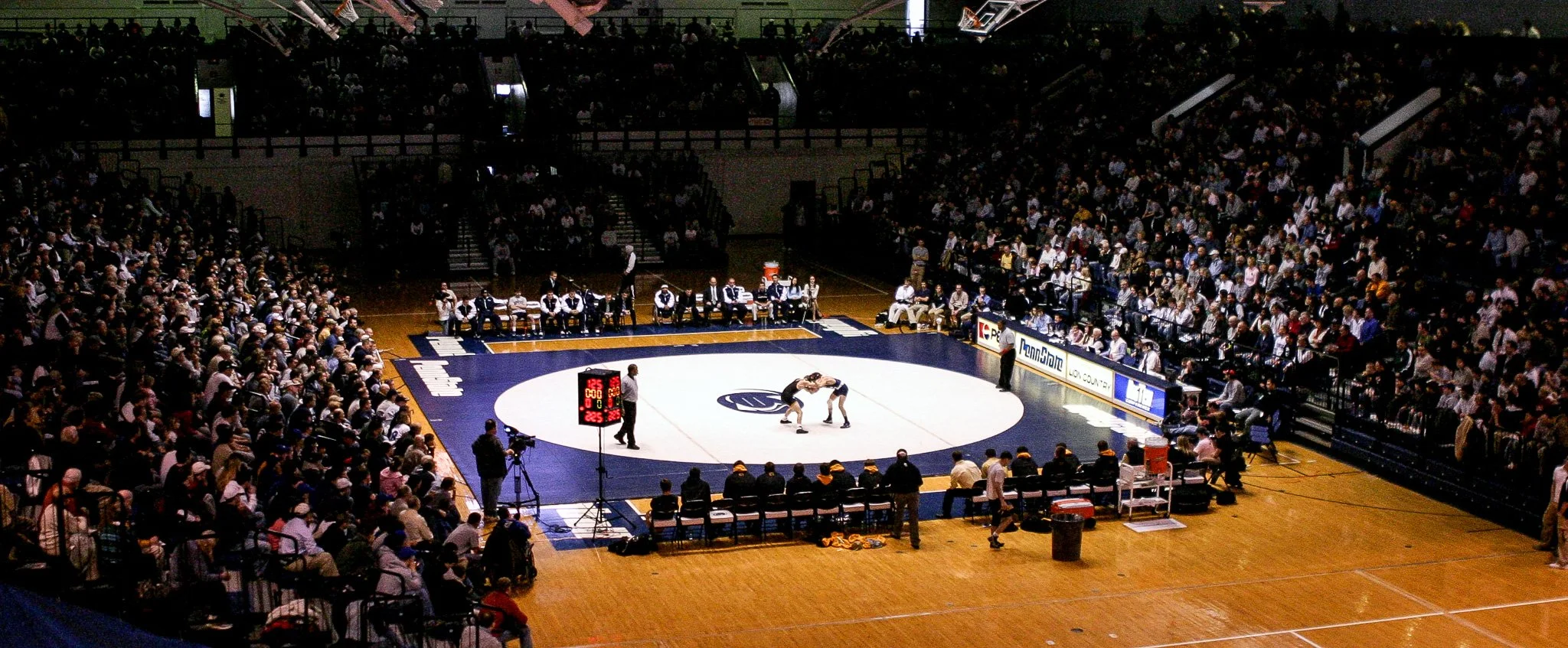 Indoor wrestling match with two wrestlers grappling on a blue and white mat surrounded by spectators and officials.