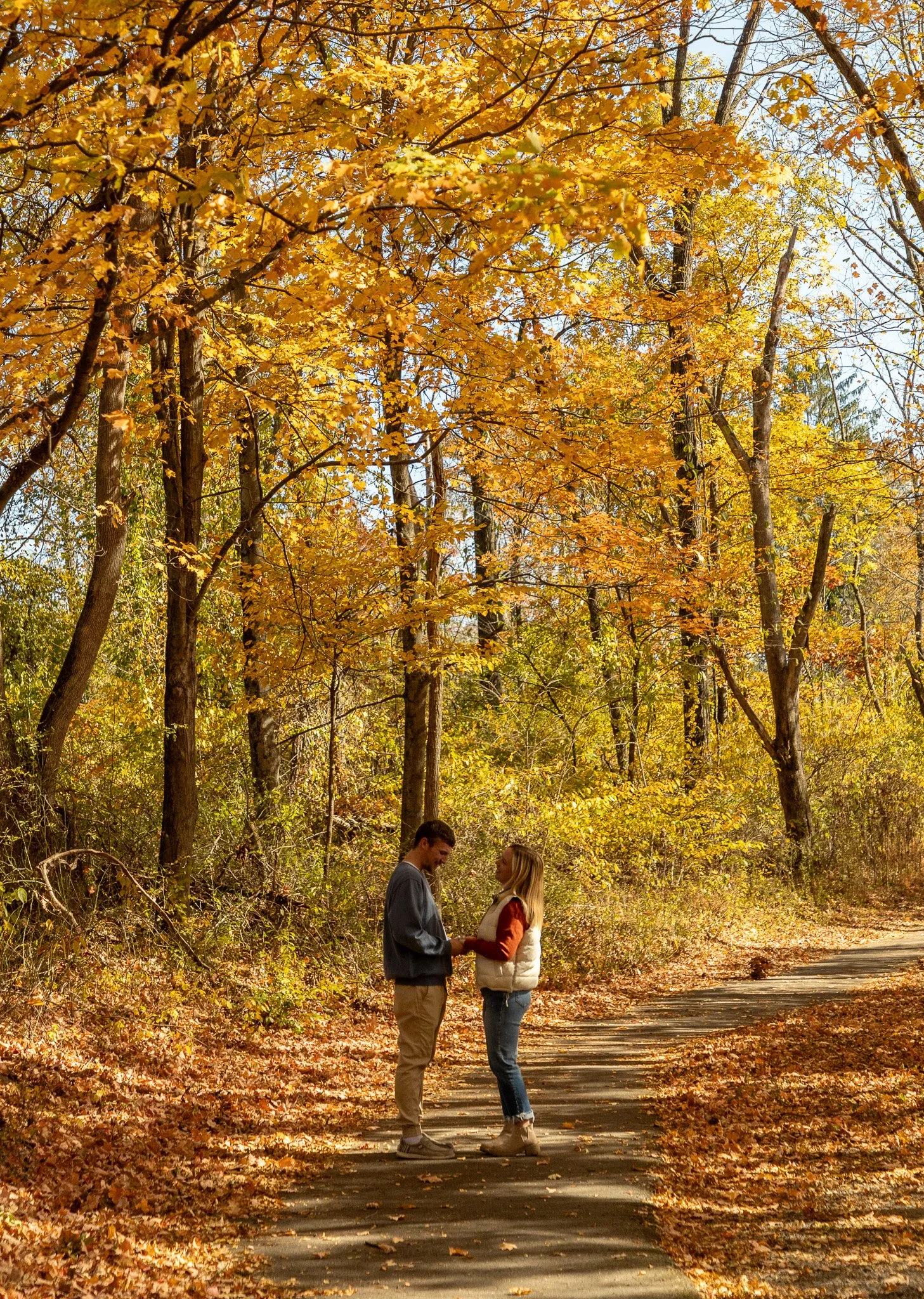 A couple standing hand in hand on a fall forest trail surrounded by yellow and orange autumn leaves.