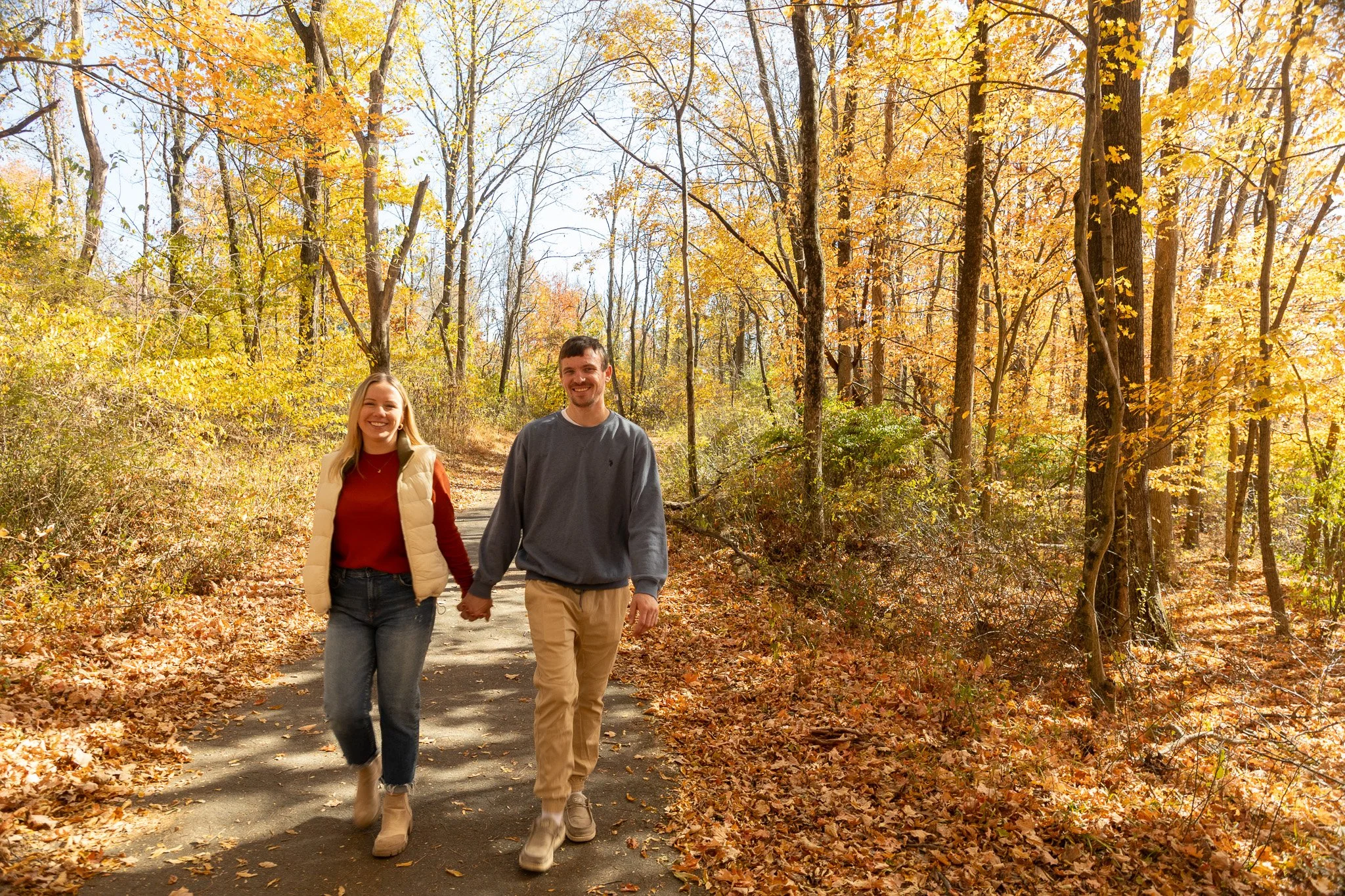 A smiling couple walking hand in hand on a paved trail through a forest with autumn-colored leaves.