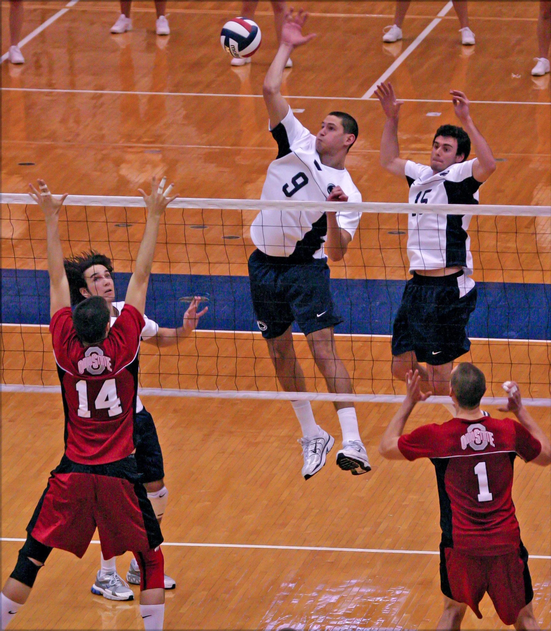 Volleyball players in action during a match, with two players jumping to spike the ball and four players on the ground preparing to block or receive.