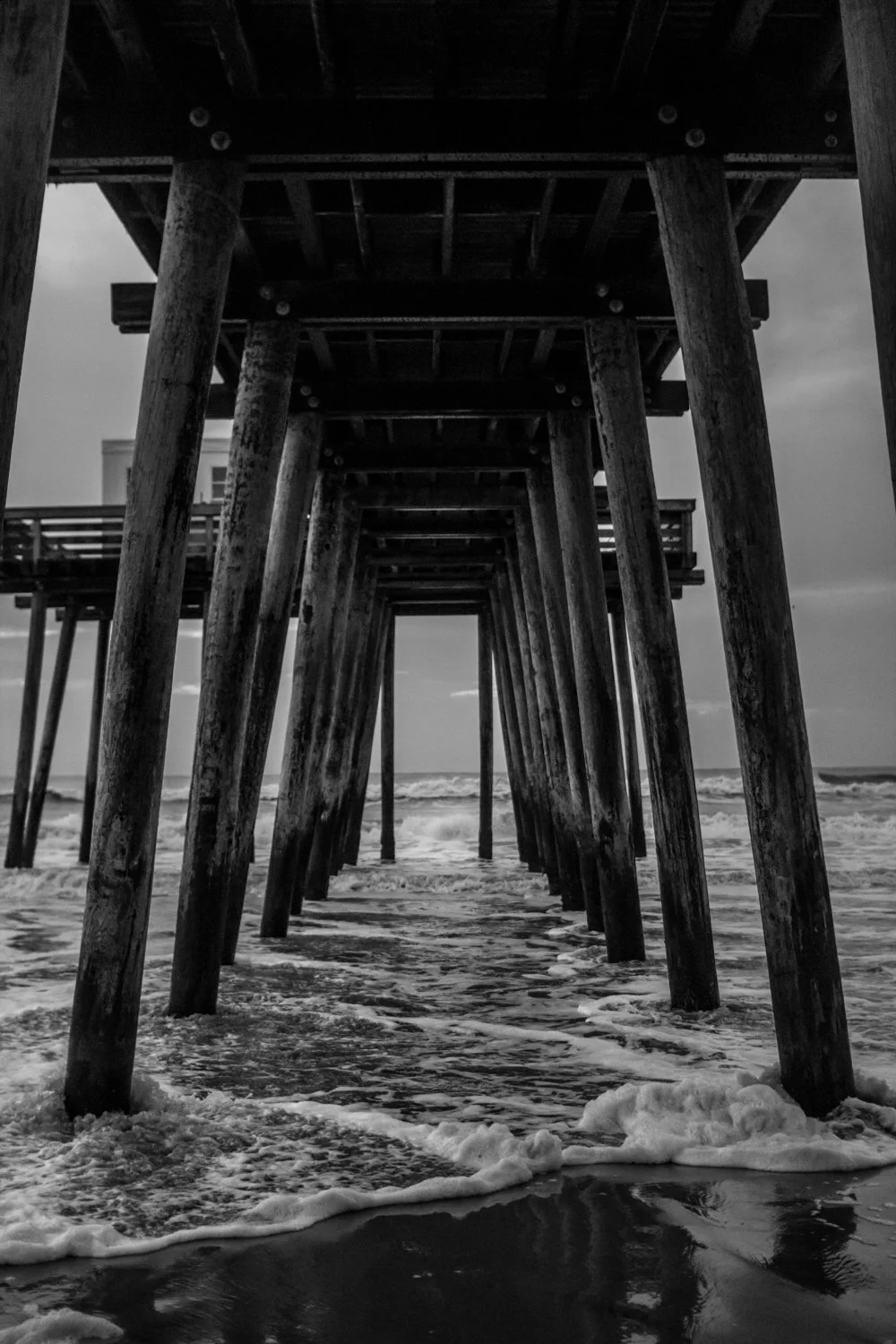 Black and white photo of a beach pier with wooden supports extending into the ocean, with waves crashing underneath.
