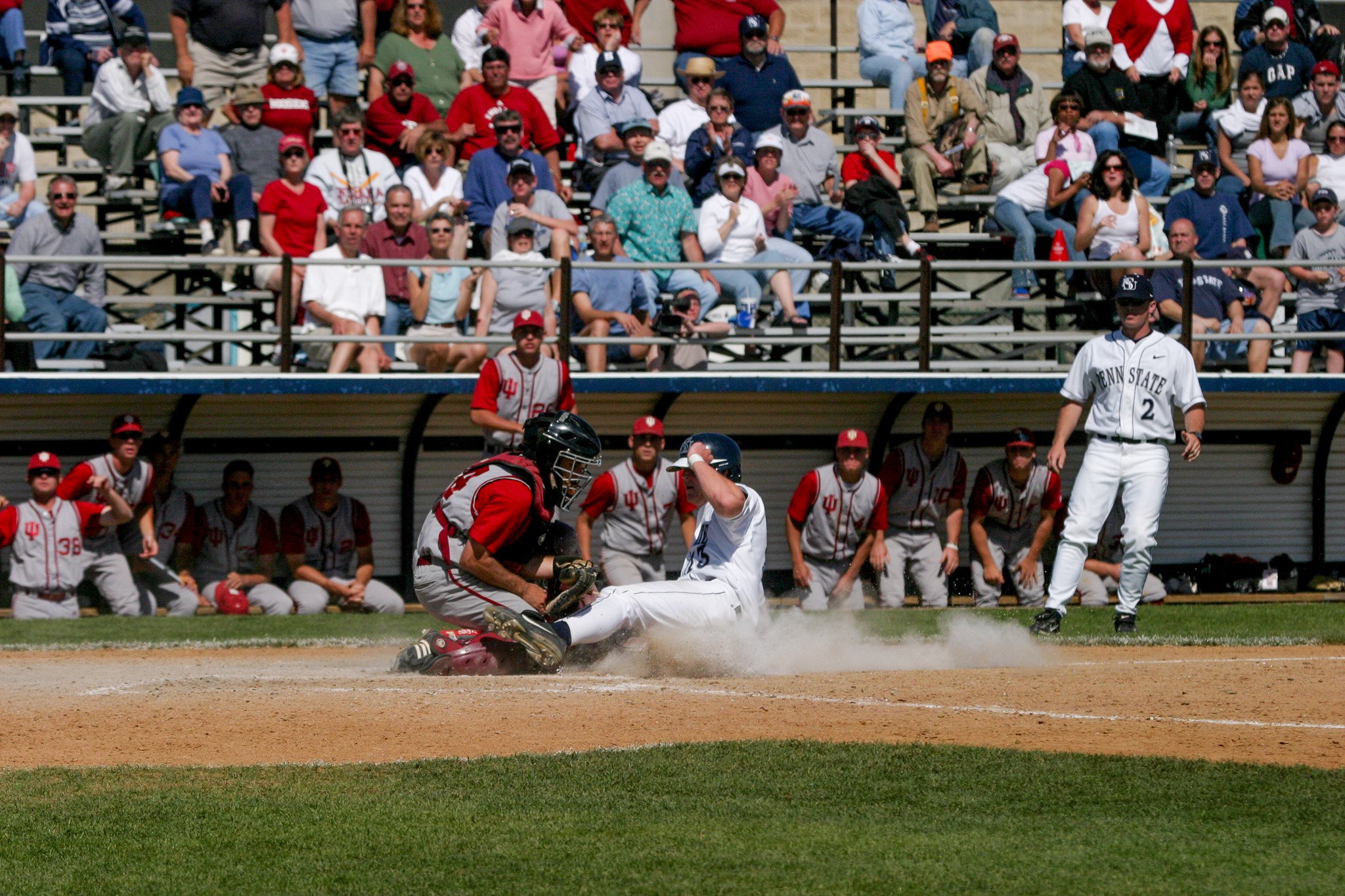 A baseball player sliding into home plate as the catcher tags him out, with spectators watching from the stands and team members on the dugout bench.